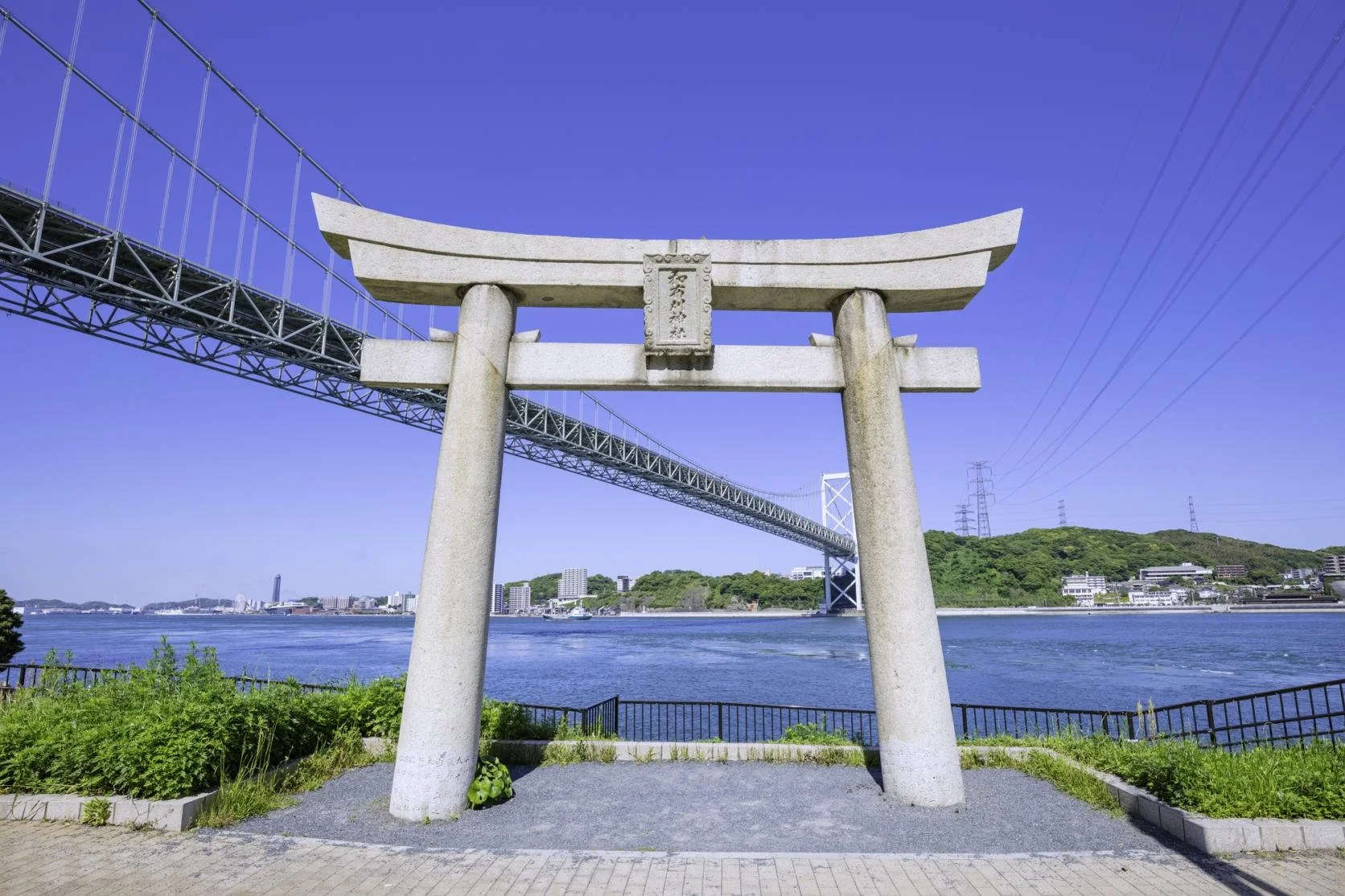Stone torii of Mekari Jinja beside the Kanmon Strait, framing the Kanmon Bridge and blue sea in Kitakyushu, Japan.