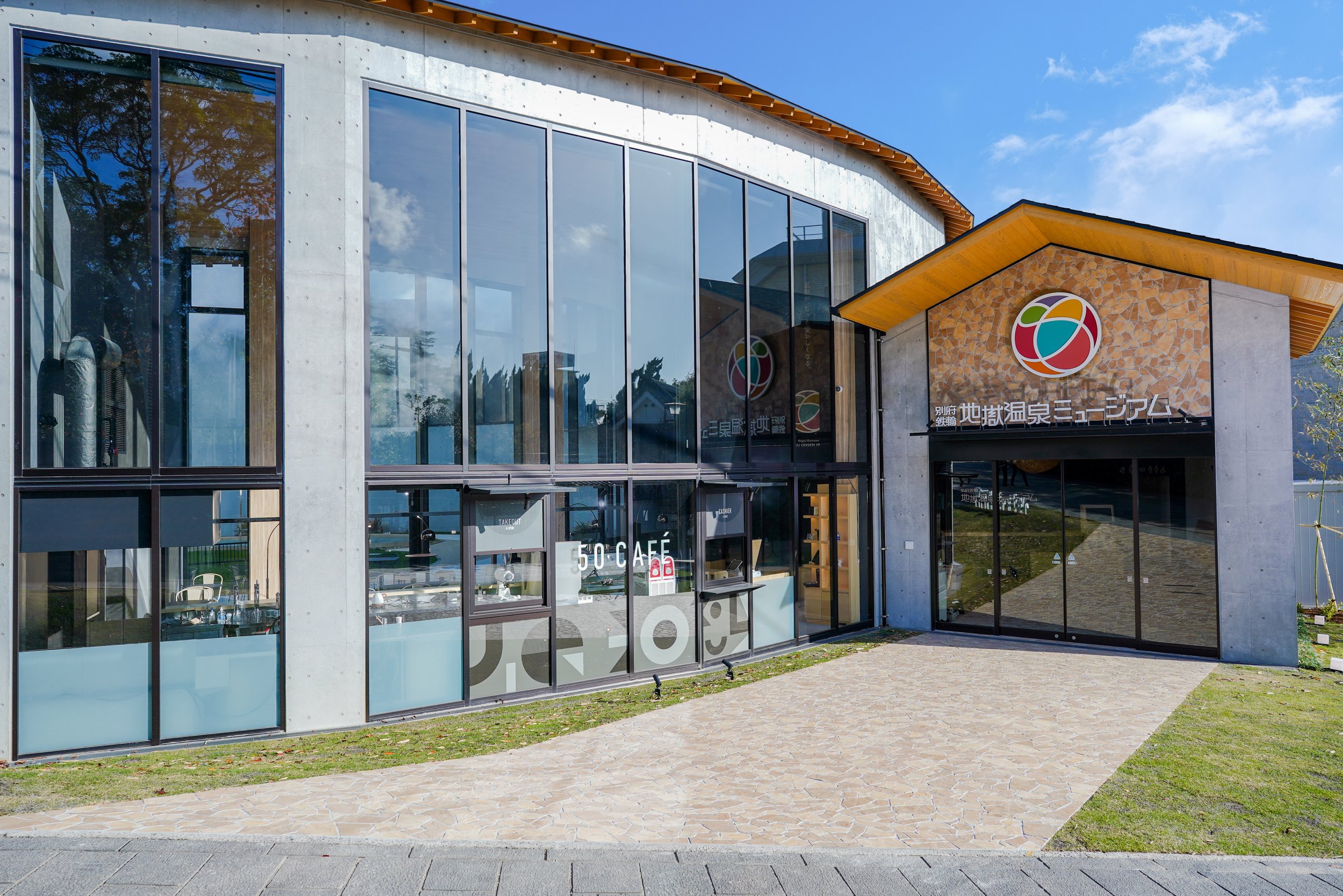 Modern glass and wood exterior of the Beppu Kannawa Jigoku Onsen Museum on a clear day, featuring its signature colorful logo and adjoining cafe.