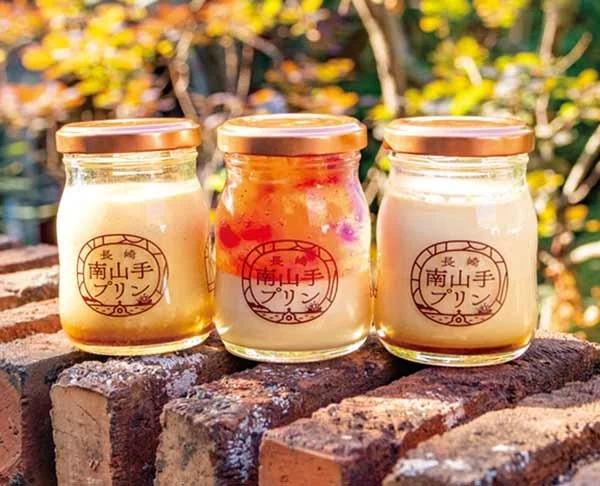 Three jars of Nagasaki Minamiyamate Pudding on a brick ledge, including the signature Stained Glass Pudding, framed by soft autumn foliage.