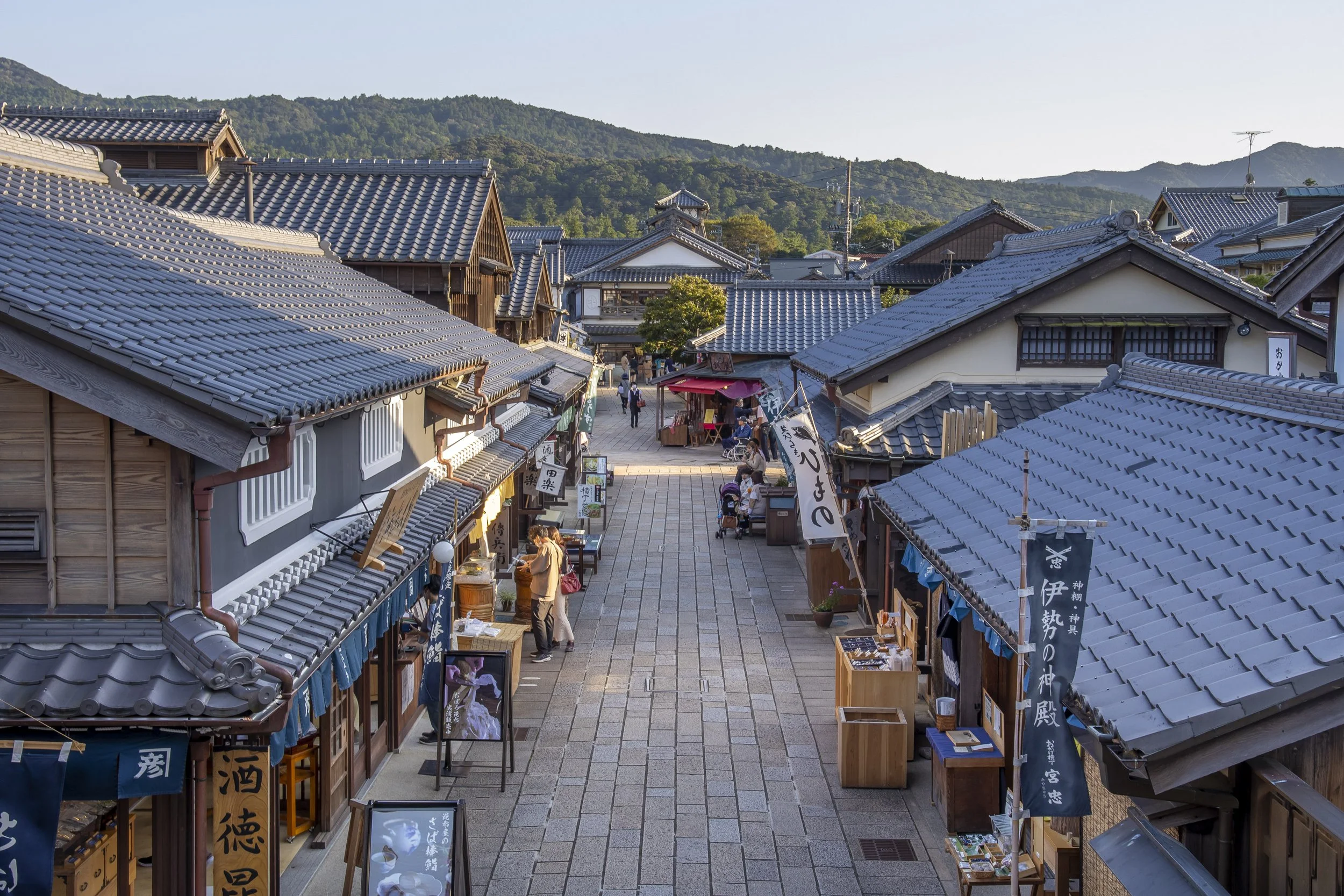 Stone-paved lane of Okage Yokocho at golden hour — Edo-era wooden shopfronts and blue noren banners line the path toward Ise Jingu's forested hills.