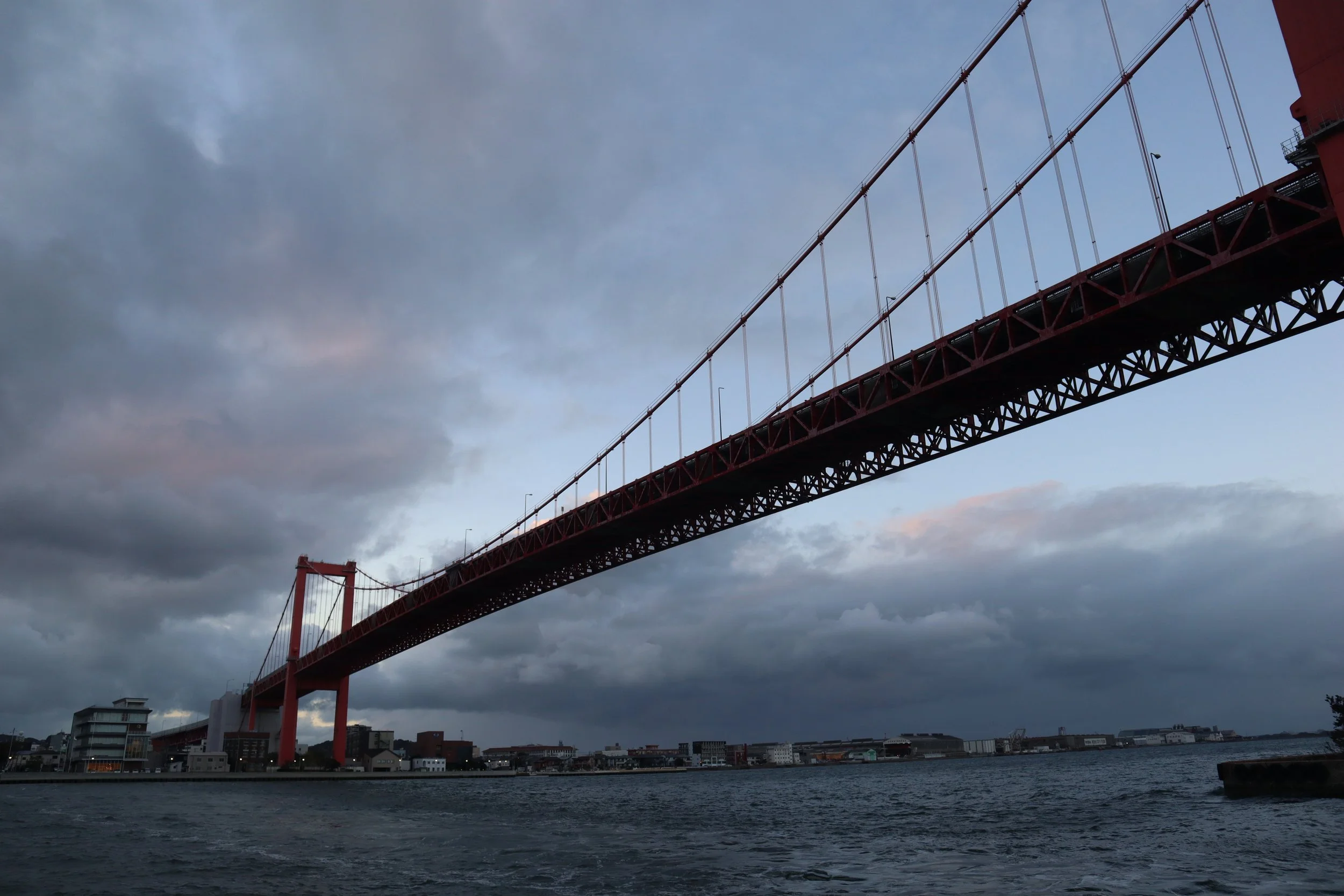 The red Wakato Bridge arcs across Dokai Bay in Kitakyushu under a moody, cloud-layered evening sky — a striking landmark of day and night.