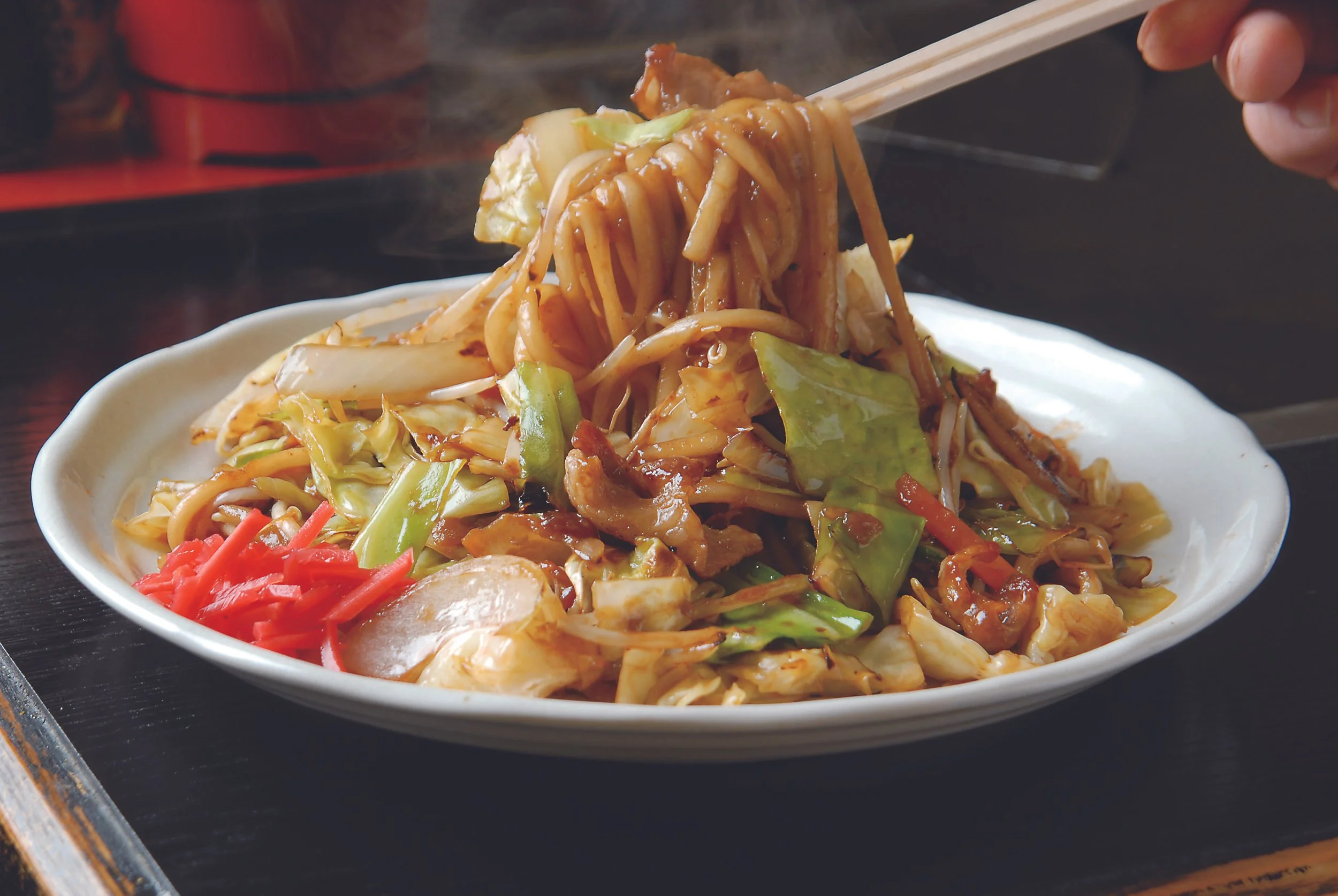 Glossy Kokura yaki-udon lifted by chopsticks — thick noodles with cabbage, pork, and pickled ginger, a postwar-born soul food of Kitakyushu.