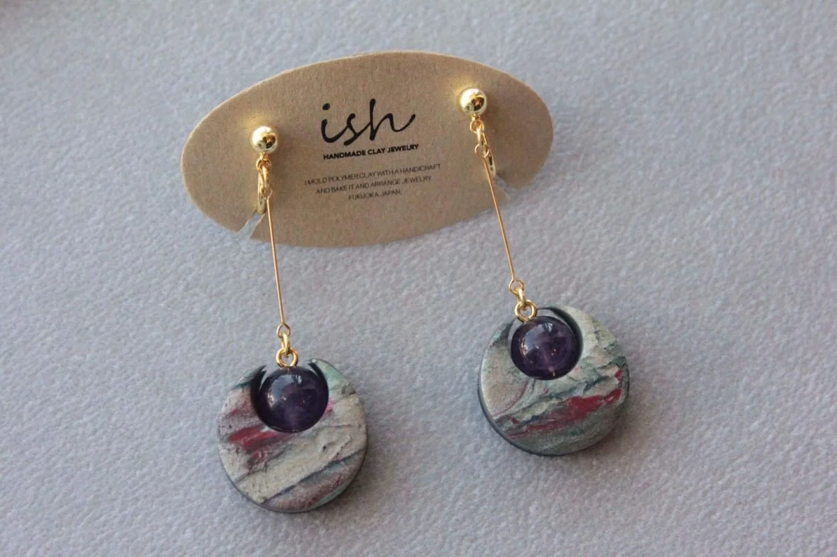 Pair of handmade clay earrings with circular bases and purple spherical beads, hanging from gold hooks, on a label reading 'ish handmade clay jewelry'.