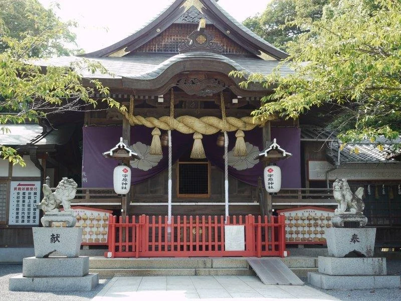 Front view of Uhara Shrine in Kanda, Fukuoka, with a massive shimenawa rope, purple noren curtains, and stone komainu guardians framing the wooden sanctuary.