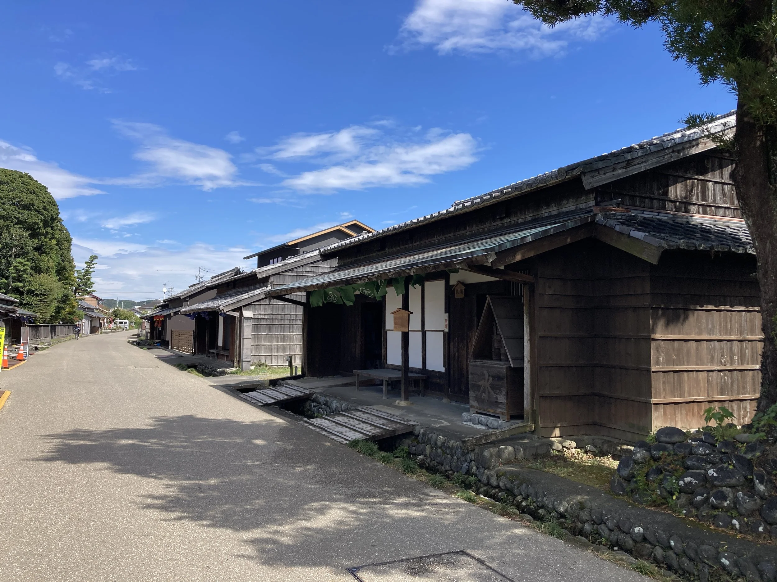 A sunlit Edo-period post town streetscape at Kawagoshi Ruins, Shimada — wooden buildings, tiled roofs, and a quiet road evoking Japan's old Tōkaidō Road.