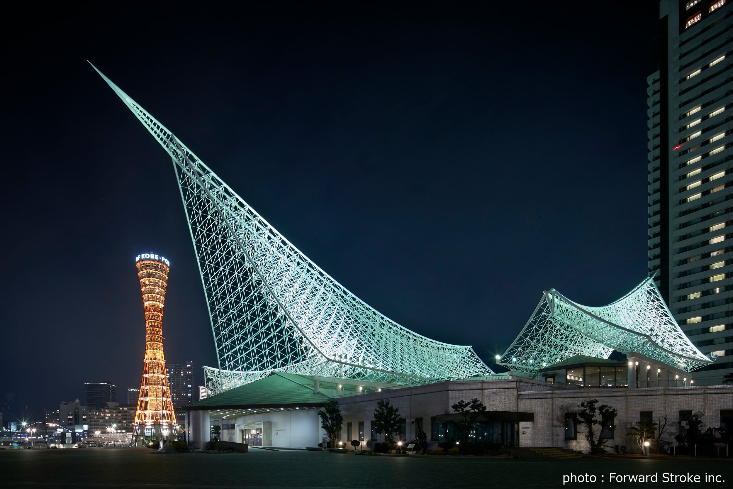 Kobe Maritime Museum's sail-shaped white lattice illuminated at night beside the glowing Kobe Port Tower in Meriken Park — an iconic Kobe waterfront landmark.