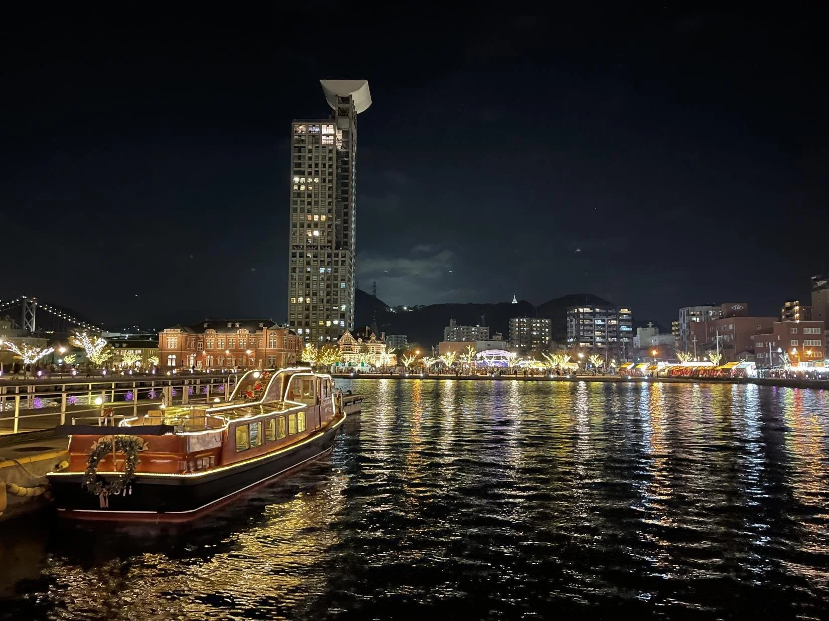 Night view of a waterfront city with illuminated buildings, decorated trees, and a boat docked at the pier.