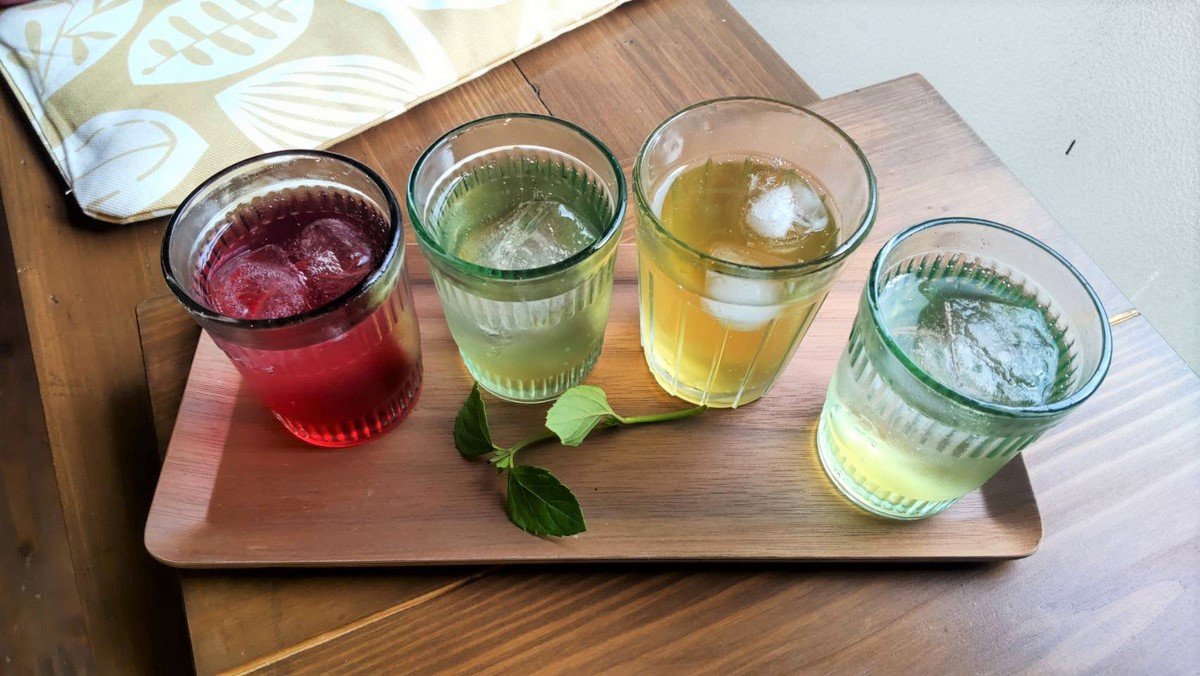 Four glasses with colored beverages and ice cubes on a wooden tray, with green leaves decoration.