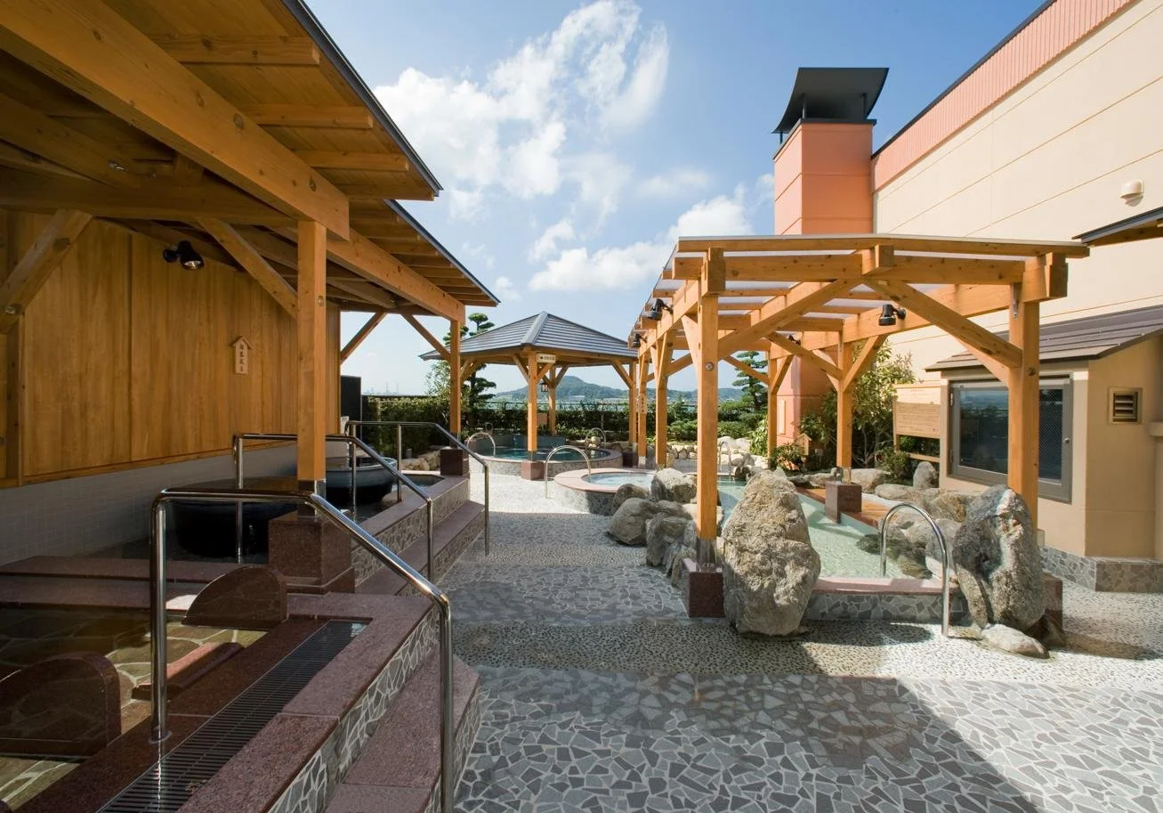 Open-air stone baths and wooden pergolas under a blue sky at Teriha Spa Resort Moji, a seaside hot spring facility overlooking the Kanmon Strait in Kitakyushu.