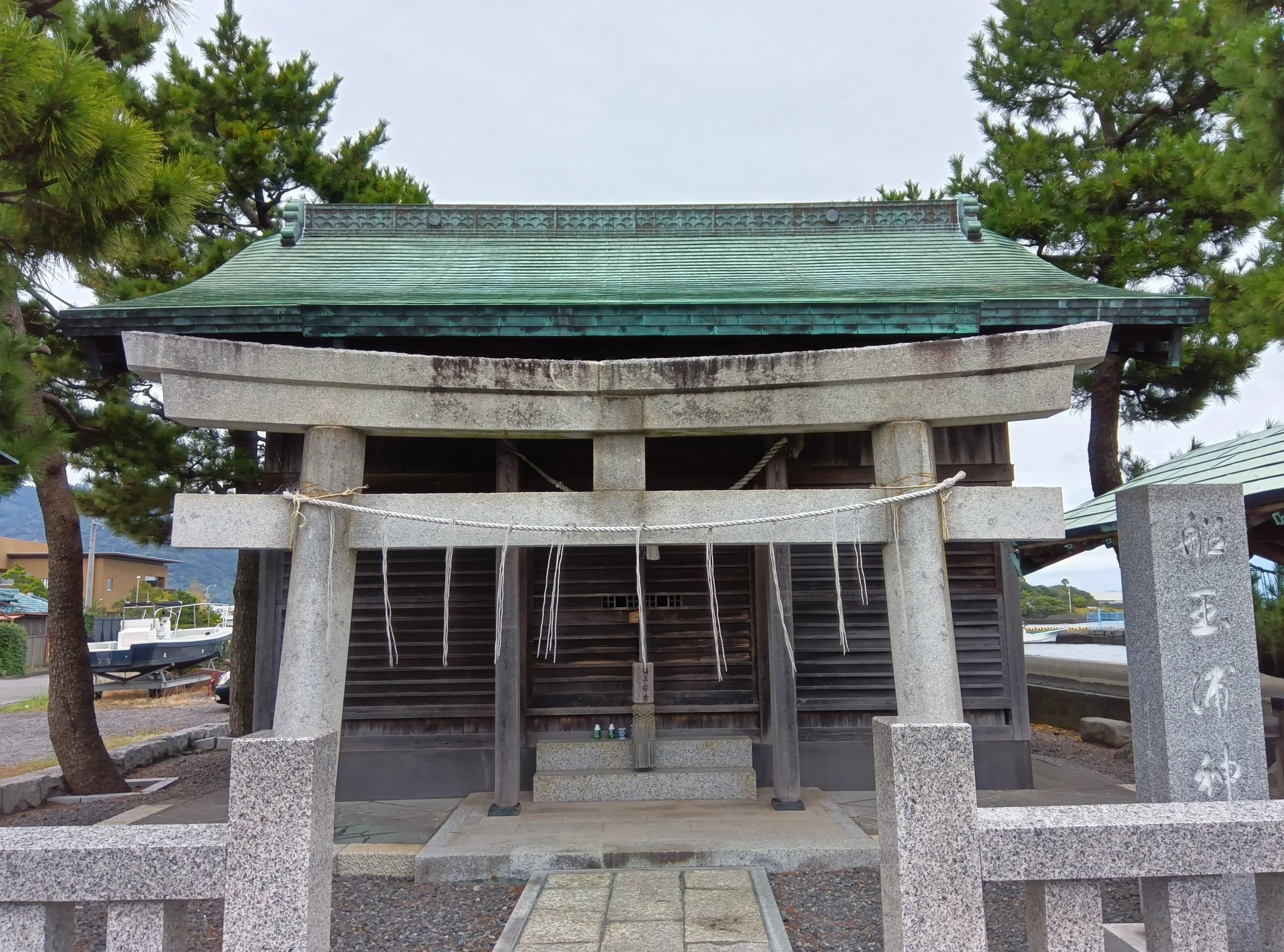 A weathered fishermen's shrine beside Yaizu Port — green-patined roof, aged shimenawa rope, and pine trees under an overcast sky.