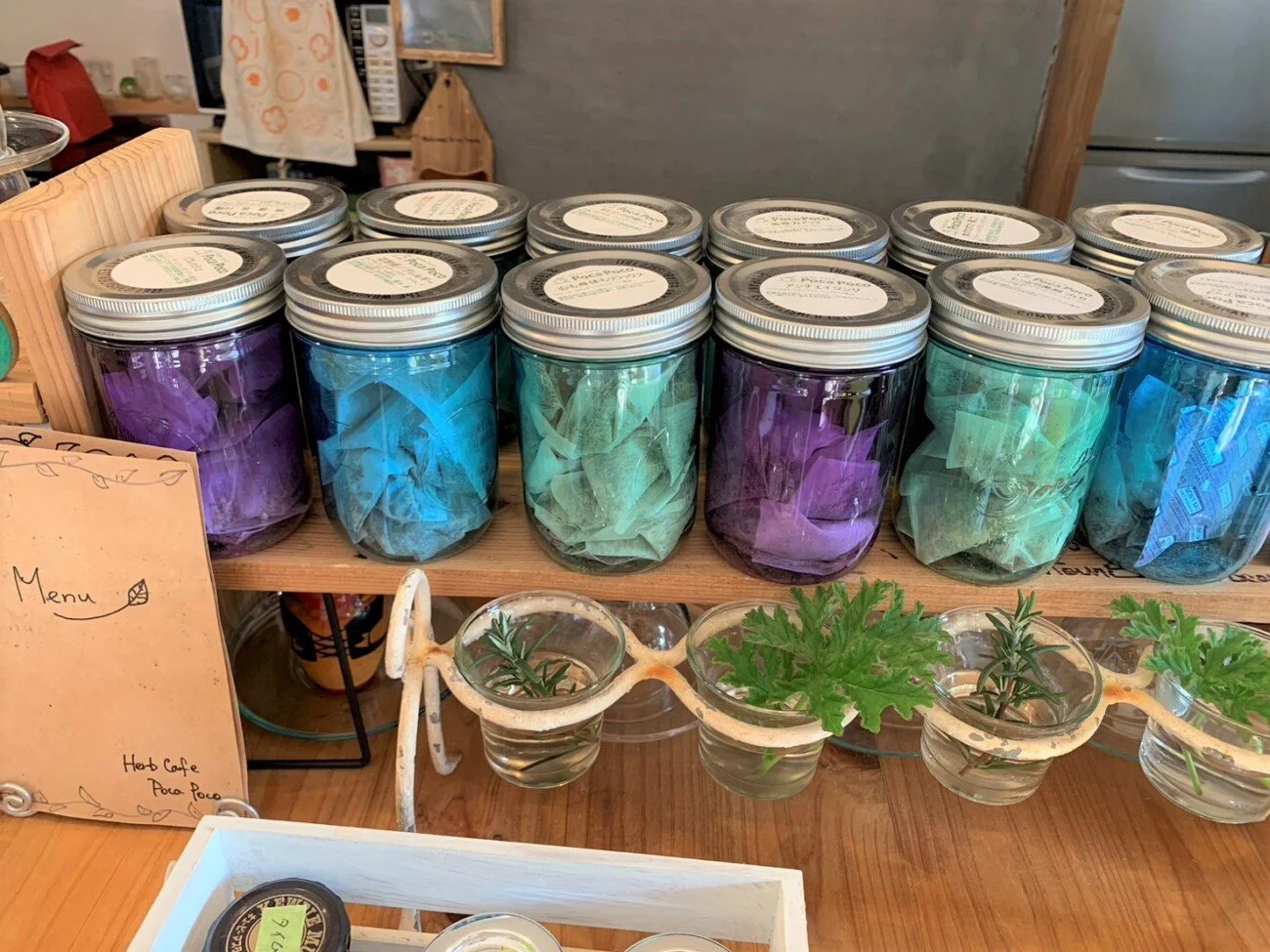 Glass jars filled with colorful tissue paper lined up on a wooden shelf, with small plant arrangements in glass cups below. A handwritten menu sign is visible on the left side.