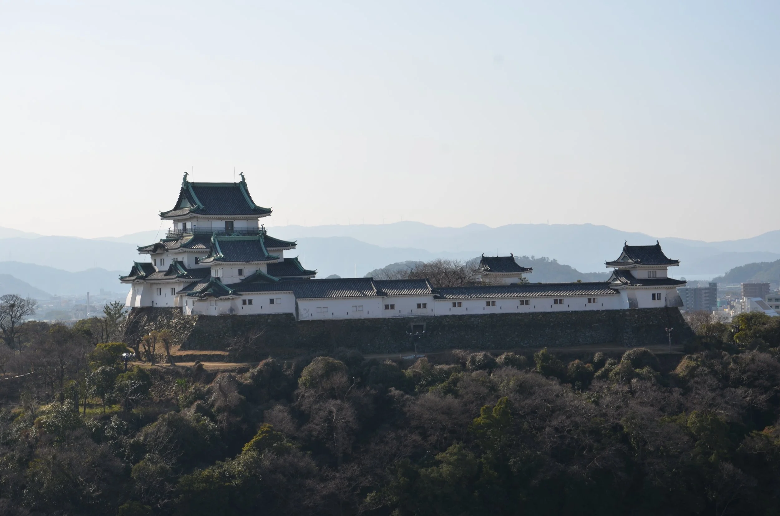 White three-tiered keep of Wakayama Castle rising above stone walls and forested Mt. Torafusu, with distant hazy mountains beyond.