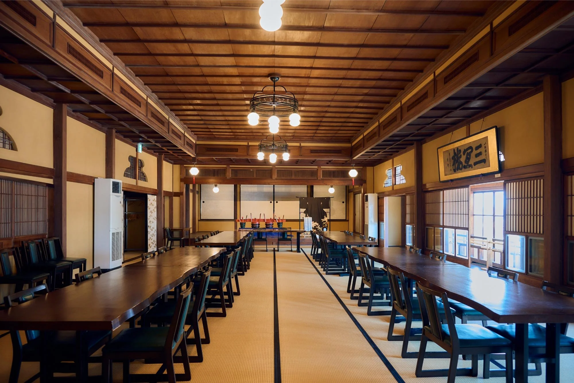Traditional Japanese large dining hall with wooden beams, long low tables, and chairs, paper lantern lighting, tatami mat flooring, and calligraphy artwork on the walls.