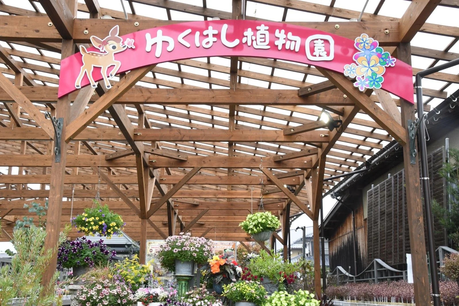 Wooden pergola with a pink 'Yukuhashi Botanical Garden' sign and colorful hanging flower baskets in Fukuoka, Japan.