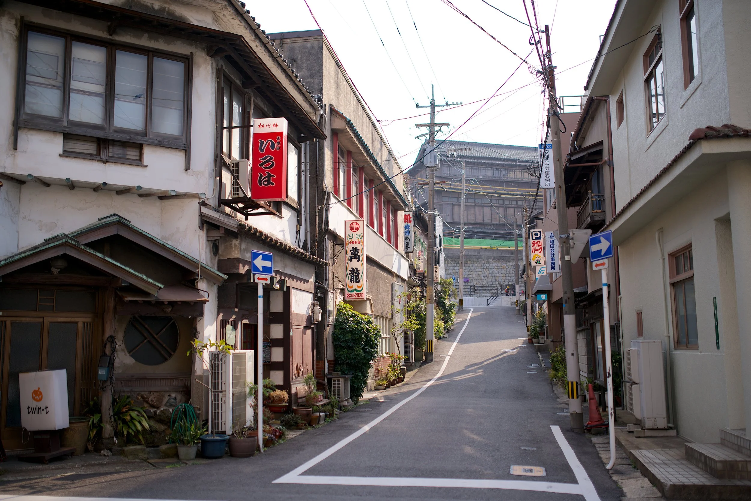 A narrow street in a Japanese neighborhood with small buildings, signs in Japanese, and overhead power lines. The street slopes uphill with residential and commercial establishments on both sides, and potted plants outside some buildings.
