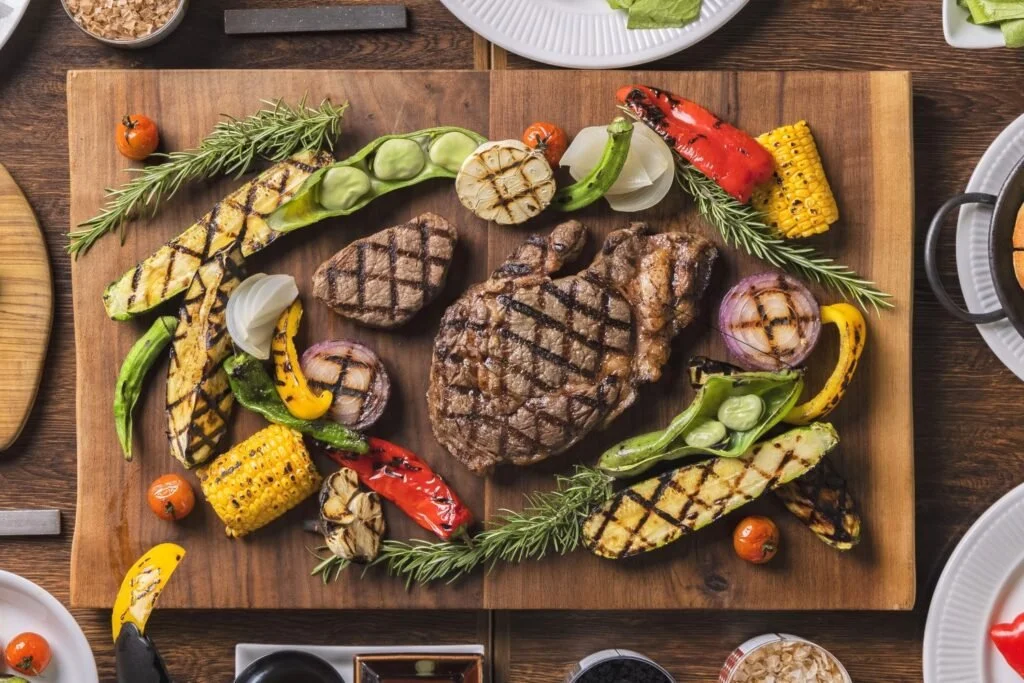Grilled steak served with grilled vegetables including corn on the cob, red and green peppers, onions, cherry tomatoes, zucchini, and grilled eggplant, garnished with sprigs of rosemary on a wooden platter.