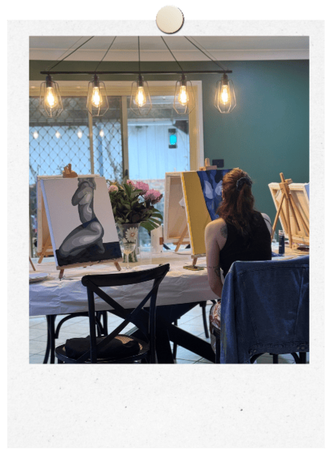 Dining table with Custom Canvases set up on table easels. Canvases are womens bodies painted in varipus shades including grey, blue and yellow. One woman sat at the table seen from behind painting a canvas. Flowers and drinks on the table.