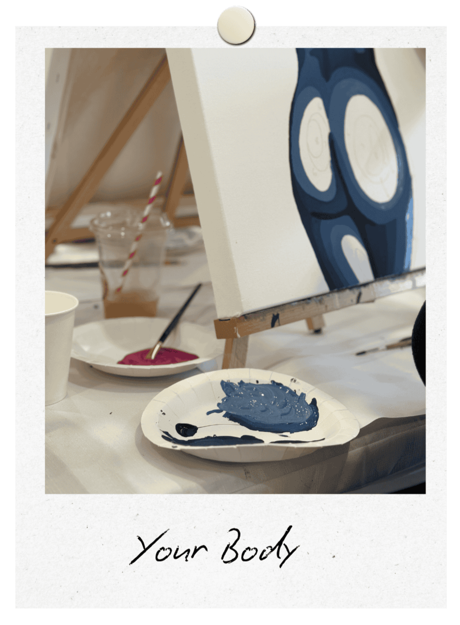 A mostly white canvas with a half painted body of a female from behind in shades of blue. Two paper plates can be seen with blue and pink paints and brushes on them.