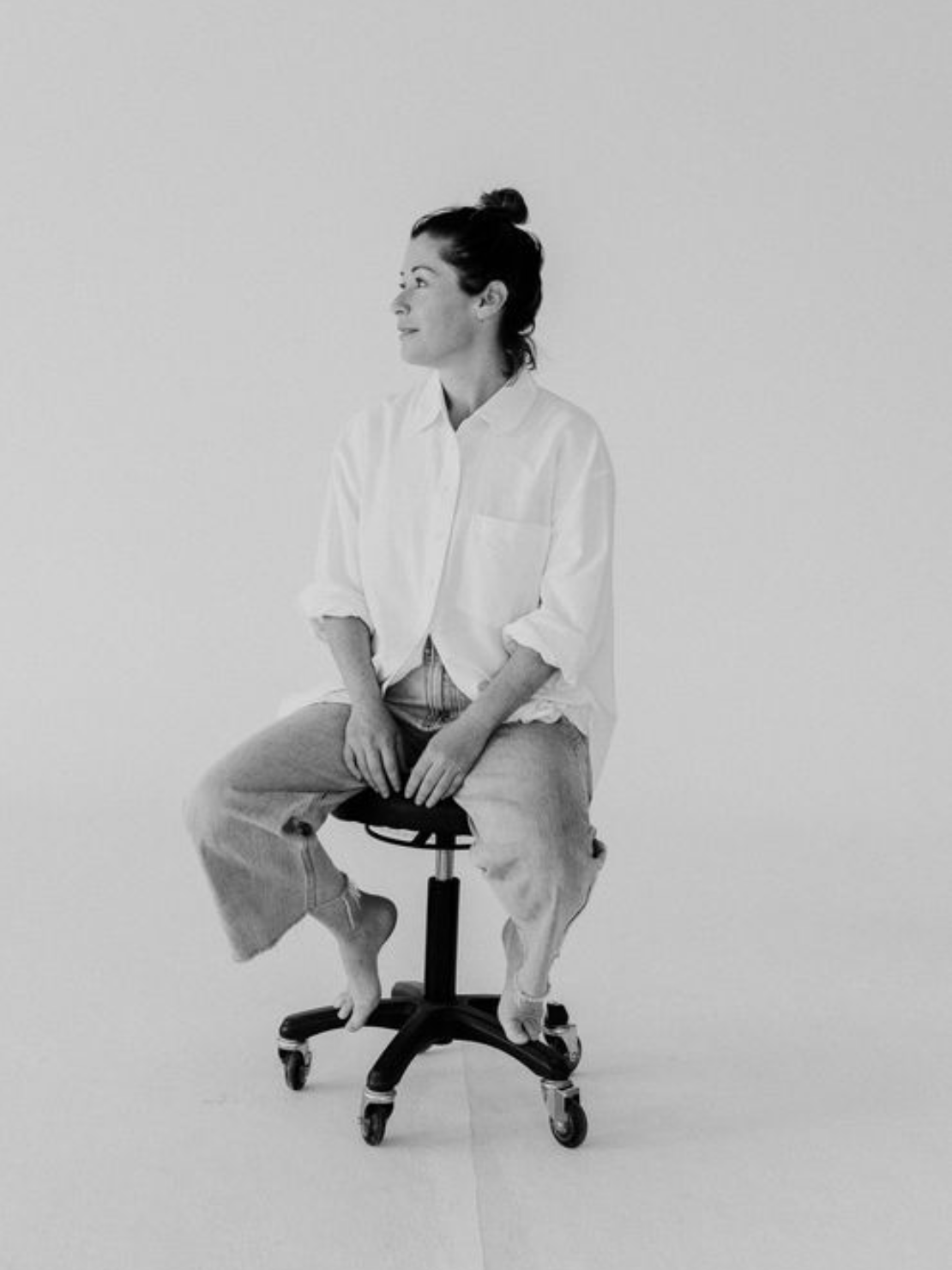 Black and white headshot of Martine Payne, a Bondi Photographer. She is in a white shirt and jeans and is sat barefoot on a stool.