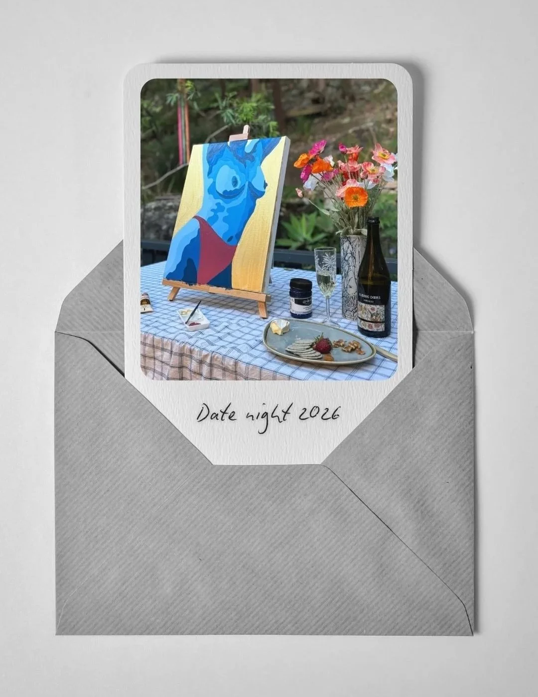 A photograph of an envelope with a photo of a table setup featuring a paint at home custom canvas, flowers, wine, and a snack, with the handwritten note 'Date night 2026' on the envelope flap.