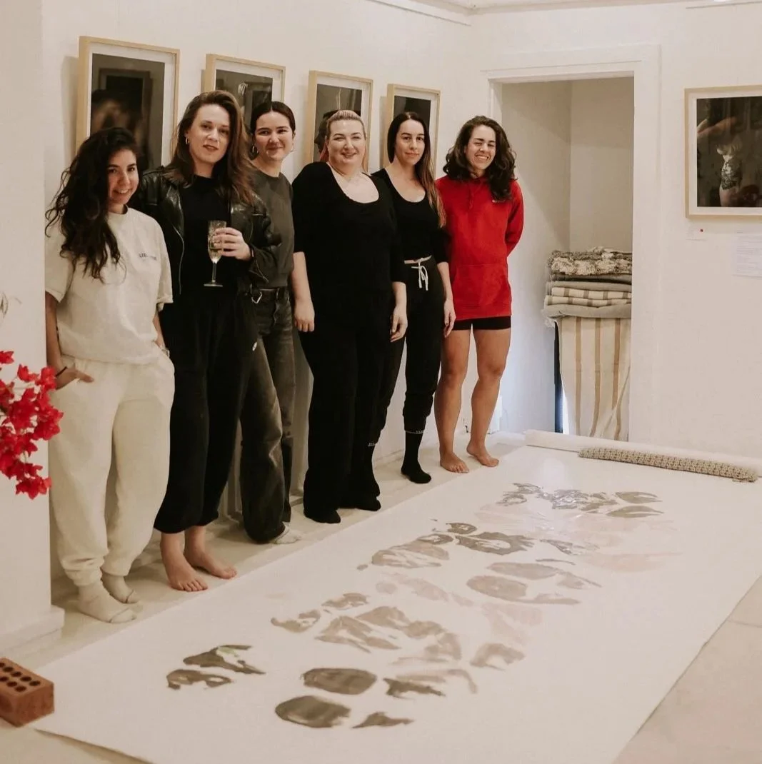 six women standing side by side in a white room with photography on the wall of female bodies. They are standing in front of a piece of art that is on a large piece of paper. it shows body prints of the women standing their in shades of nude