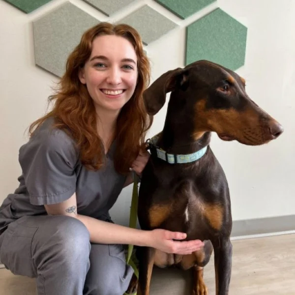 A woman in medical scrubs smiling and sitting next to a large Doberman dog in an indoor setting with decorative wall panels.