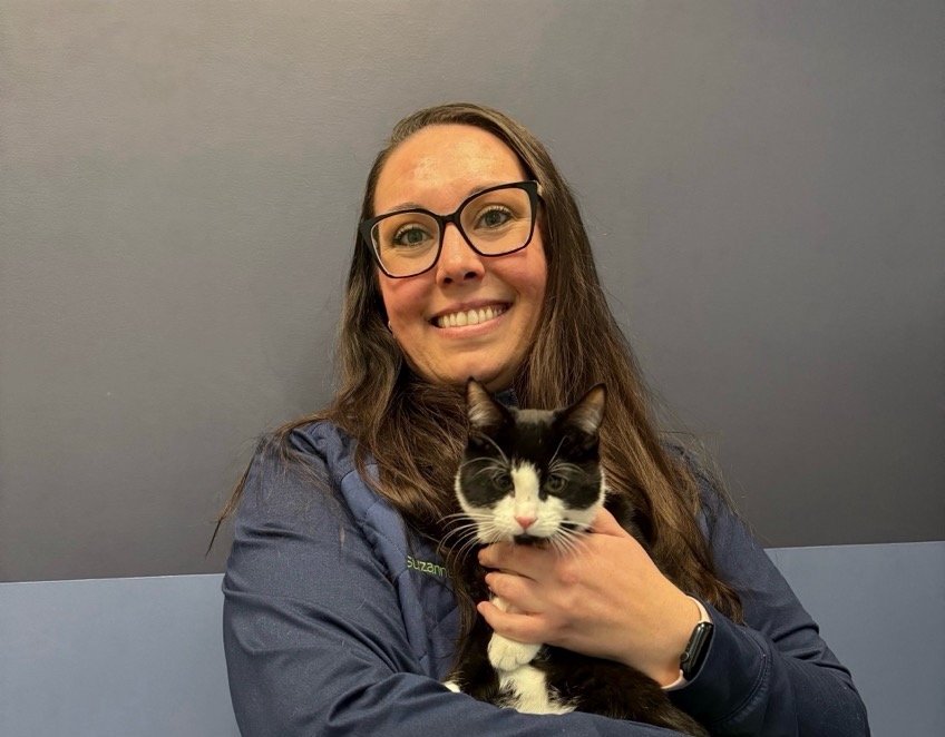 A woman with long brown hair wearing glasses and a dark jacket, smiling and holding a black and white cat in front of a plain gray wall.