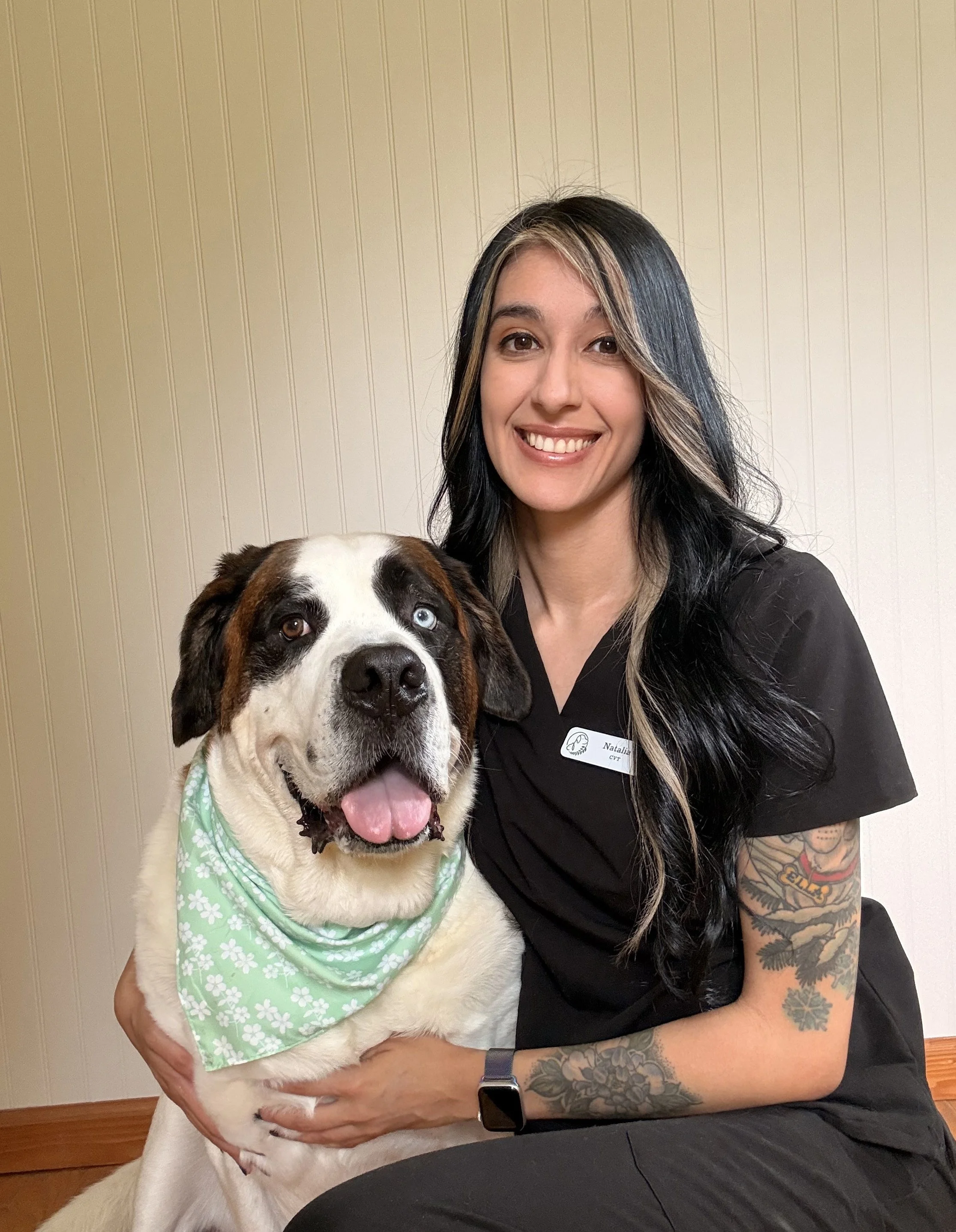 A woman with long black hair and tattoos on her arm, wearing a black shirt with a name tag, smiling while holding a large dog with one eye blue and one eye brown, wearing a green bandana.