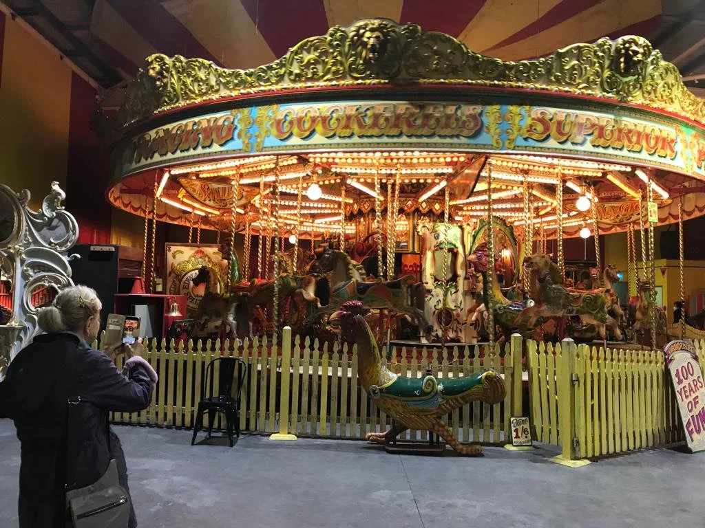 Vintage carousel with colorful horses and lights, person taking a photo, yellow picket fence, historical fairground atmosphere.