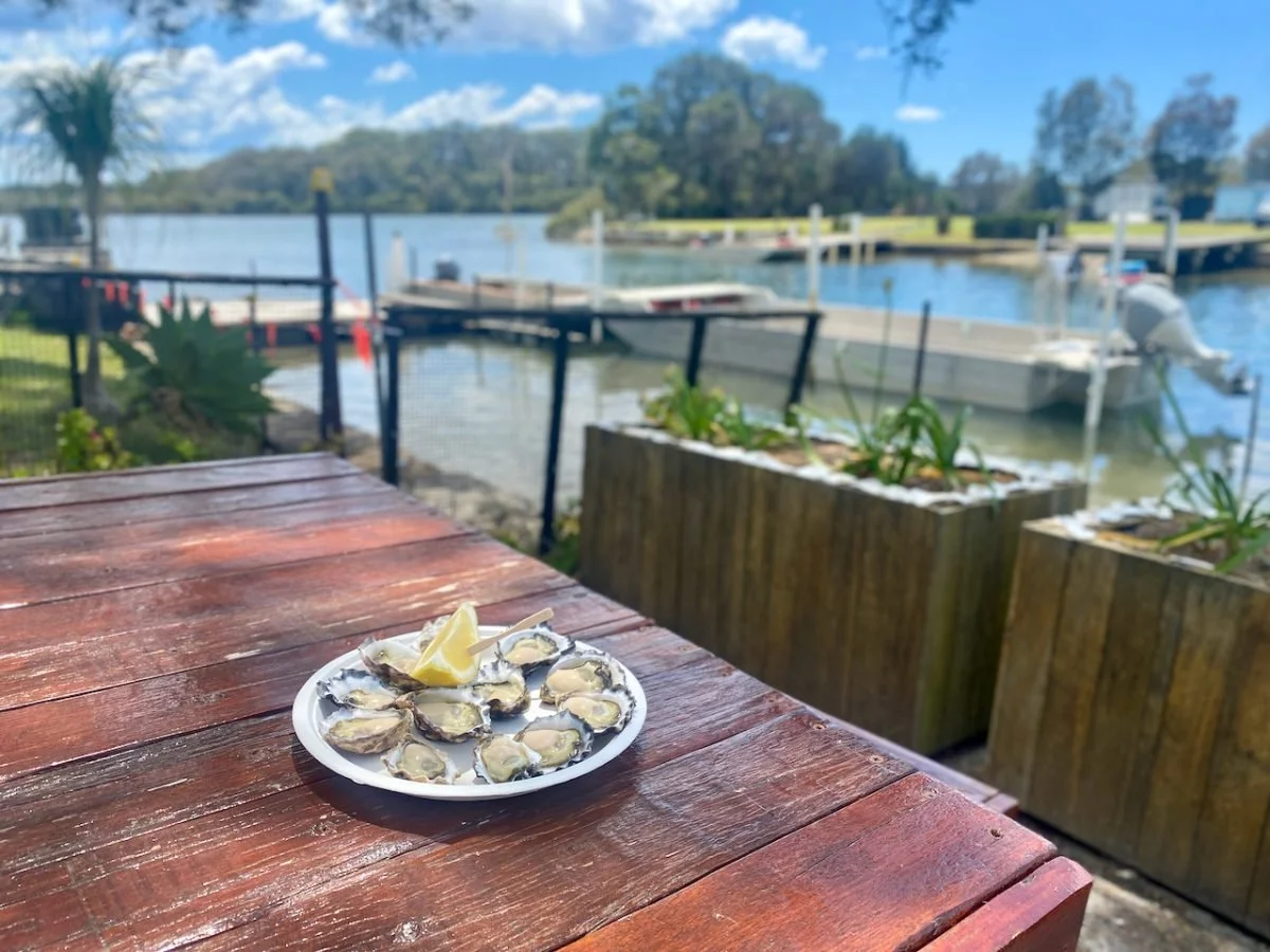 Plate of oysters with lemon on wooden table by waterfront with docks and boats.