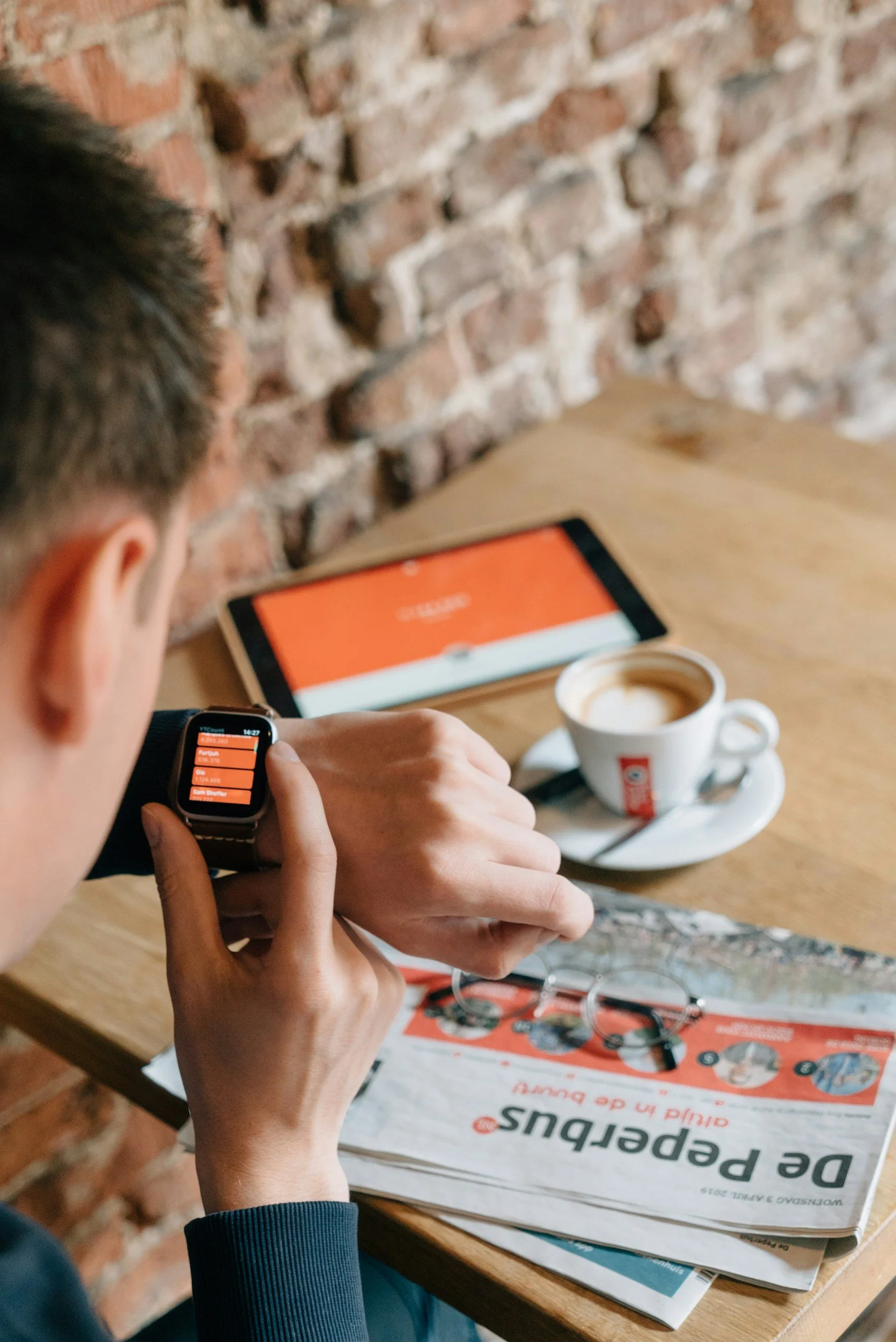 Uomo che controlla un orologio intelligente con un menu di caffè, con una tazza di cappuccino e occhiali su un tavolo di legno, contro una parete di mattoni.