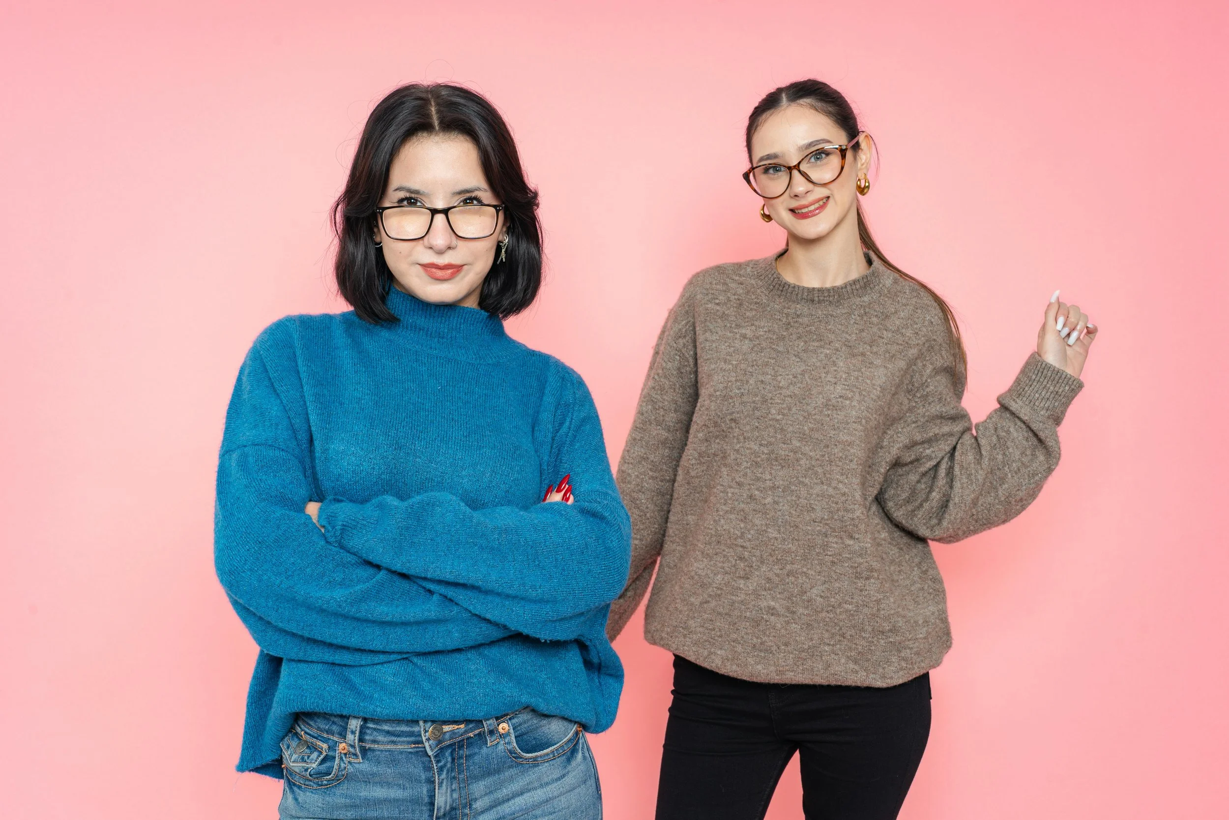 Due giovani donne con occhiali, una indossa un maglione blu e jeans, l'altra un maglione marrone e pantaloni neri, posano contro uno sfondo rosa.
