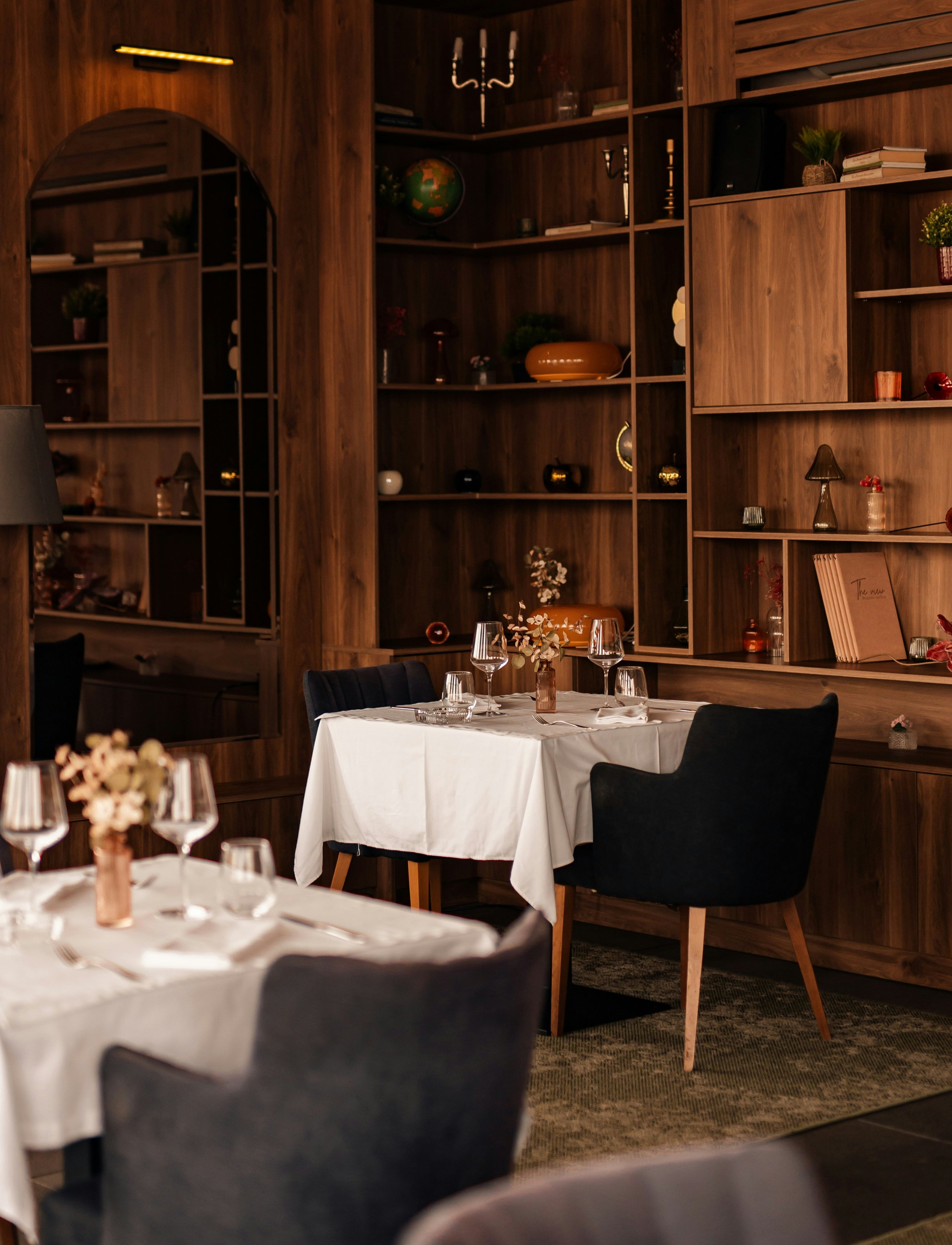 Elegant restaurant dining room with wood-paneled shelves, decorative objects, set tables, and upholstered chairs