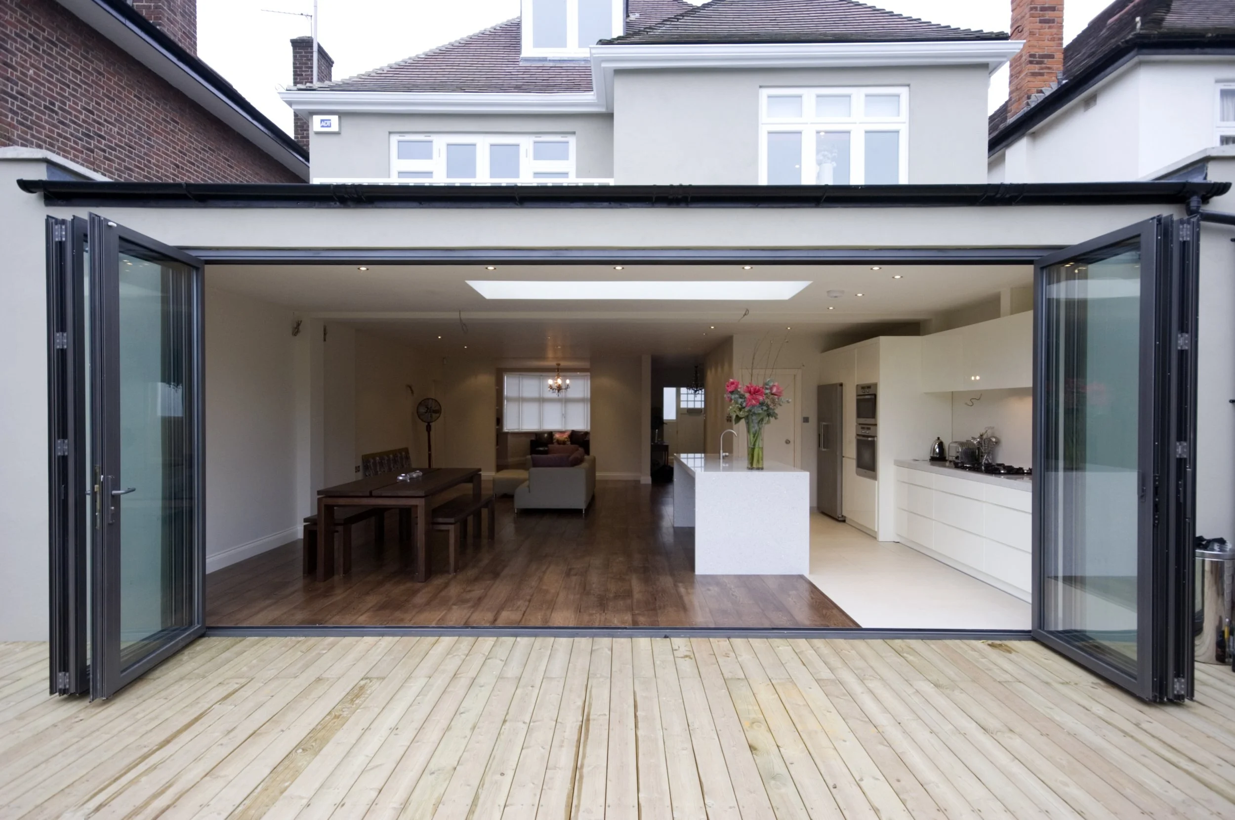 Modern home extension with open folding glass doors, wood deck, white kitchen, dining area, and living room.