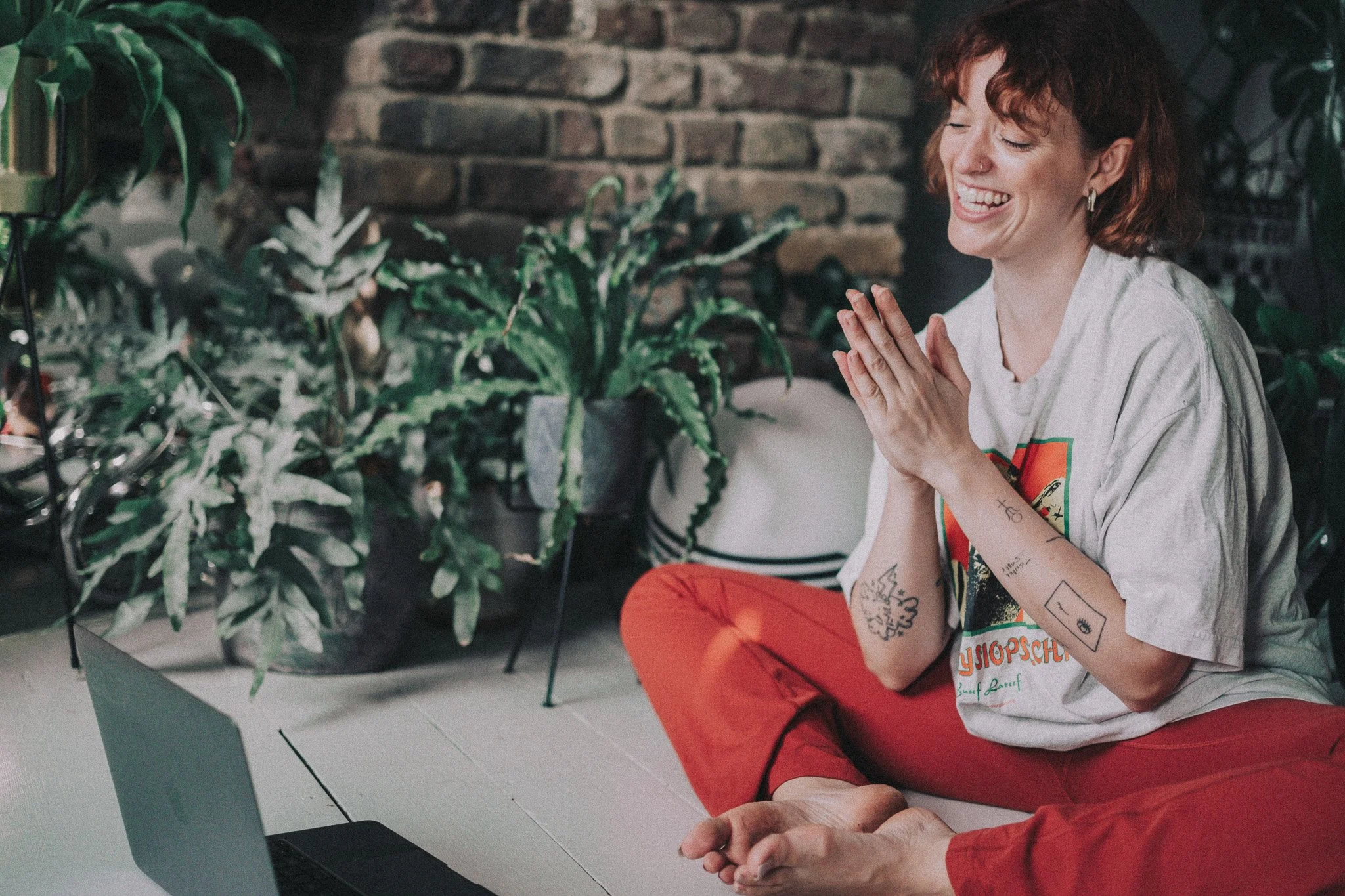 A woman with tattoos on her arms and short reddish hair, smiling and sitting cross-legged on the floor, in front of a laptop, with plants and a brick wall in the background.