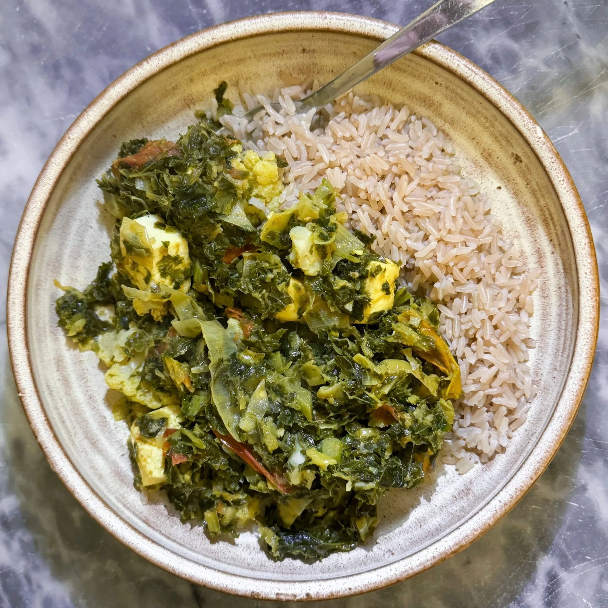A bowl of cooked rice and a green vegetable stew, possibly collard greens, on a metal surface.