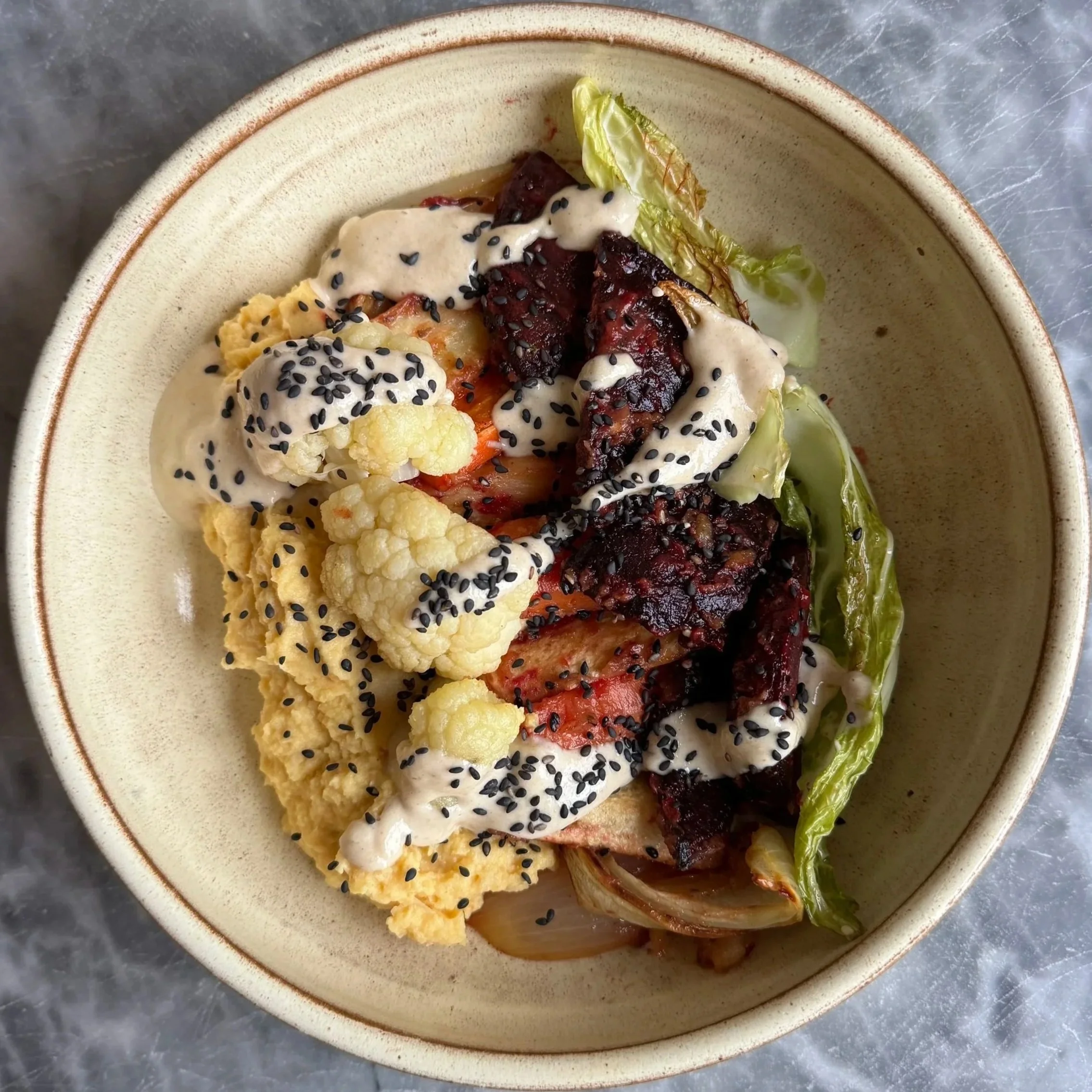 A bowl of ramen noodles topped with cauliflower, leafy lettuce, sliced tomatoes, grilled meat, black sesame seeds, and drizzle of white sauce.
