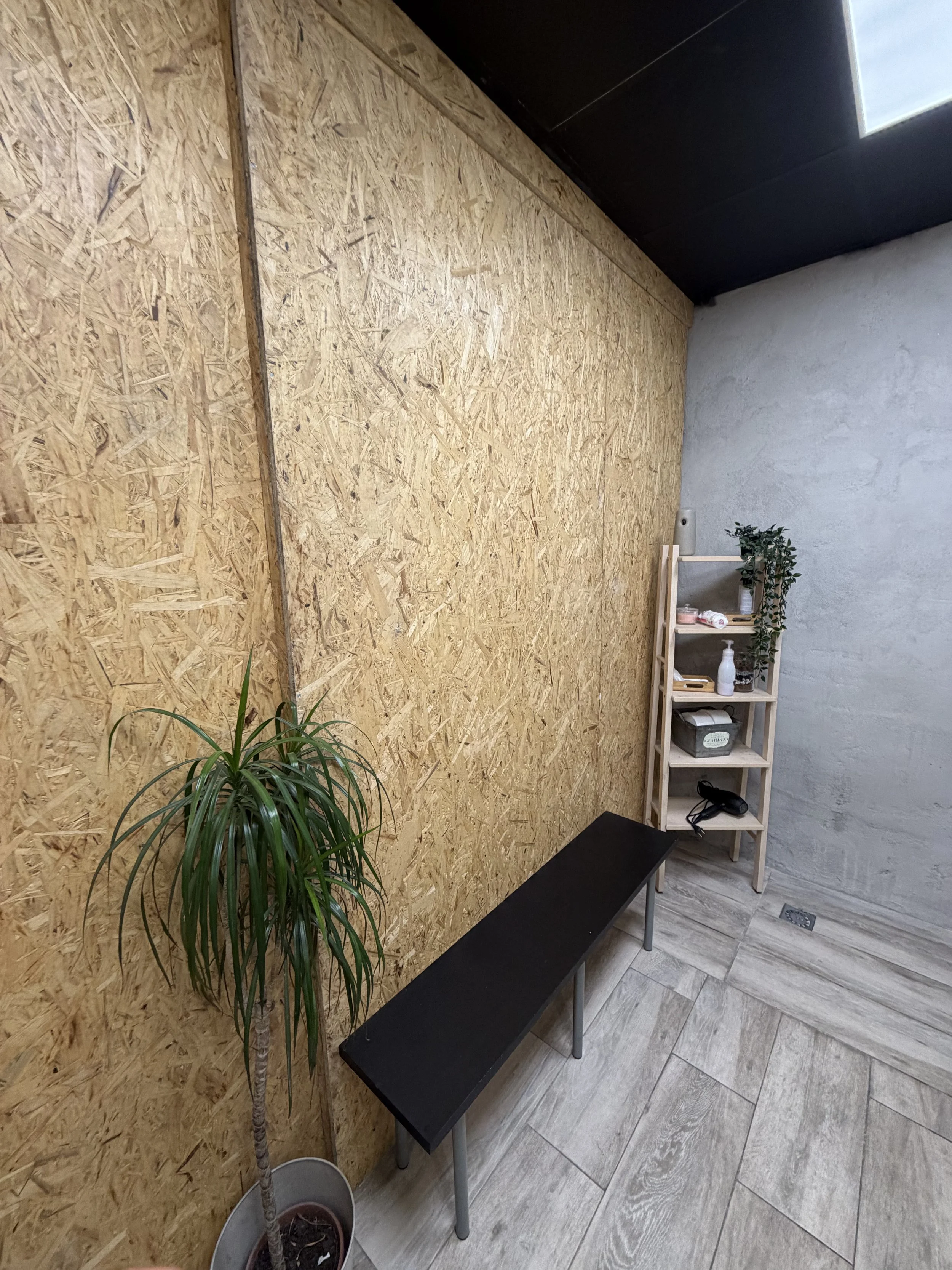 Espacio interior con pared de madera contrachapada, planta en maceta, banco negro, y estantería de madera con objetos decorativos y de oficina en el suelo de baldosas de madera.