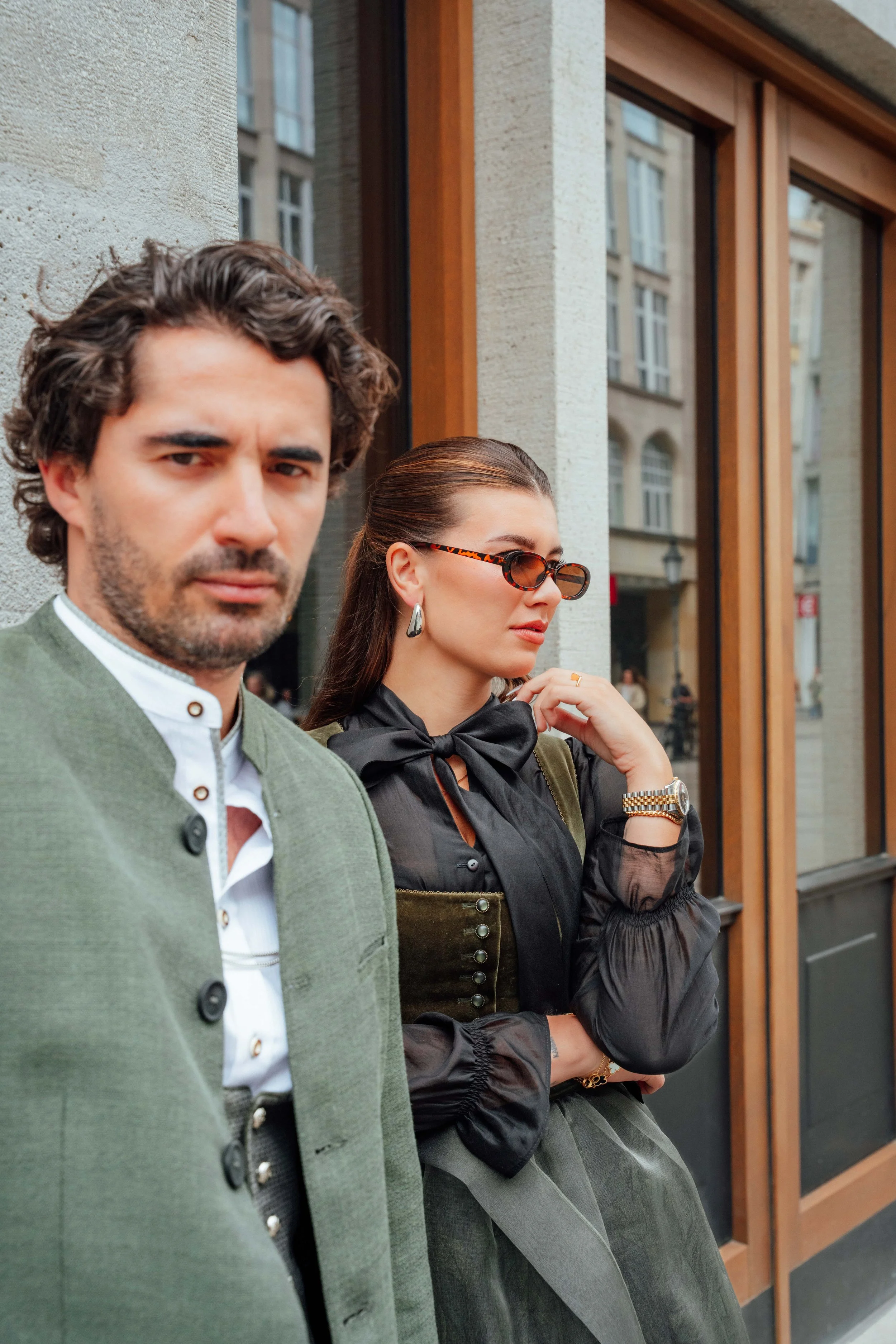 Ein Mann mit dunklen, lockigen Haaren und einem grünen Jacke steht vor einer Ziegelmauer. Dahinter eine Frau mit langen dunklen Haaren, Sonnenbrille, schwarzen Bluse, Brokatrock und Schmuck, die an einer Fensterfront in einer Stadt steht.