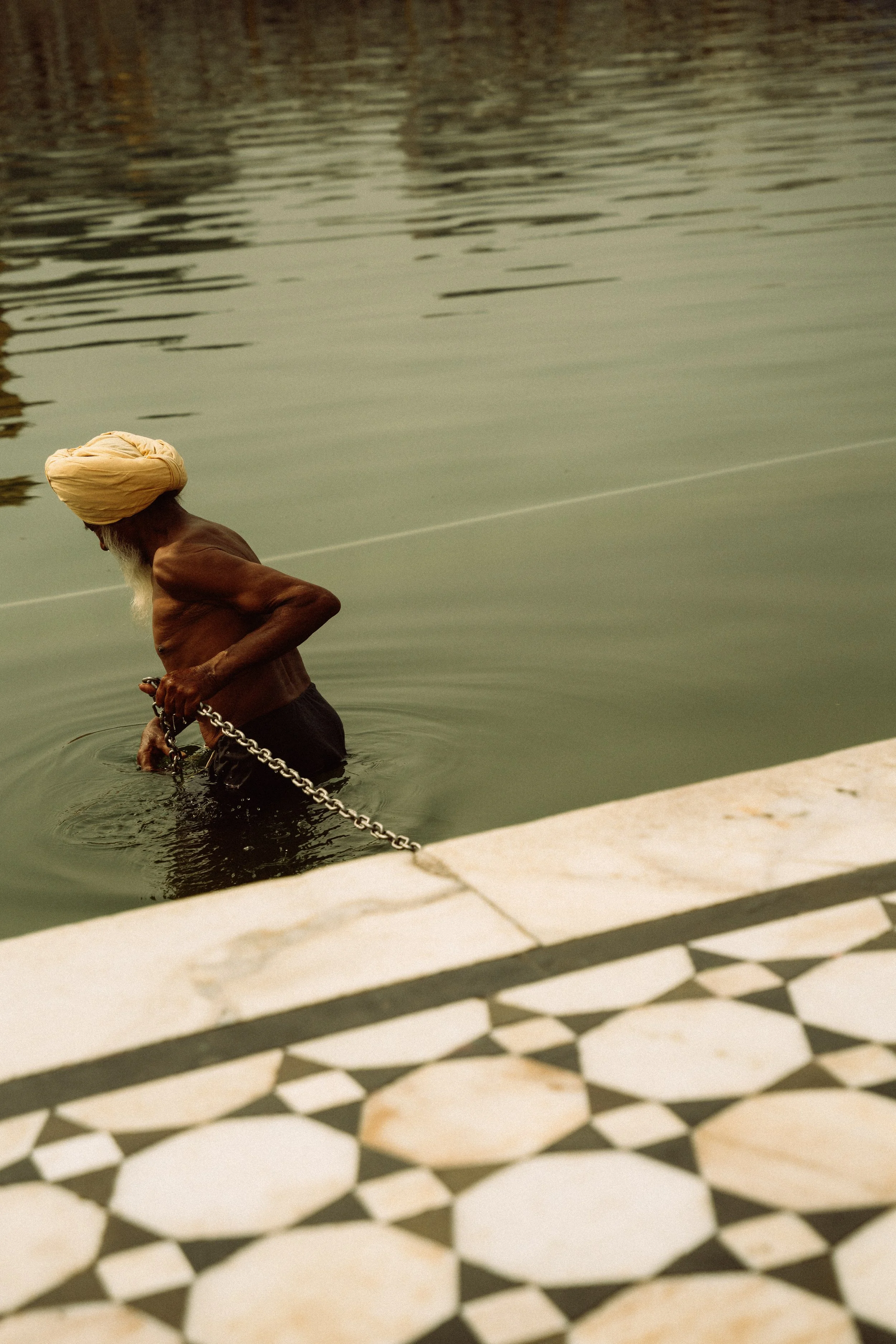 Älterer Mann mit Turban und dunkler Hose, der an der Wasserlinie steht und an einer Kette zieht bei Sonnenuntergang.