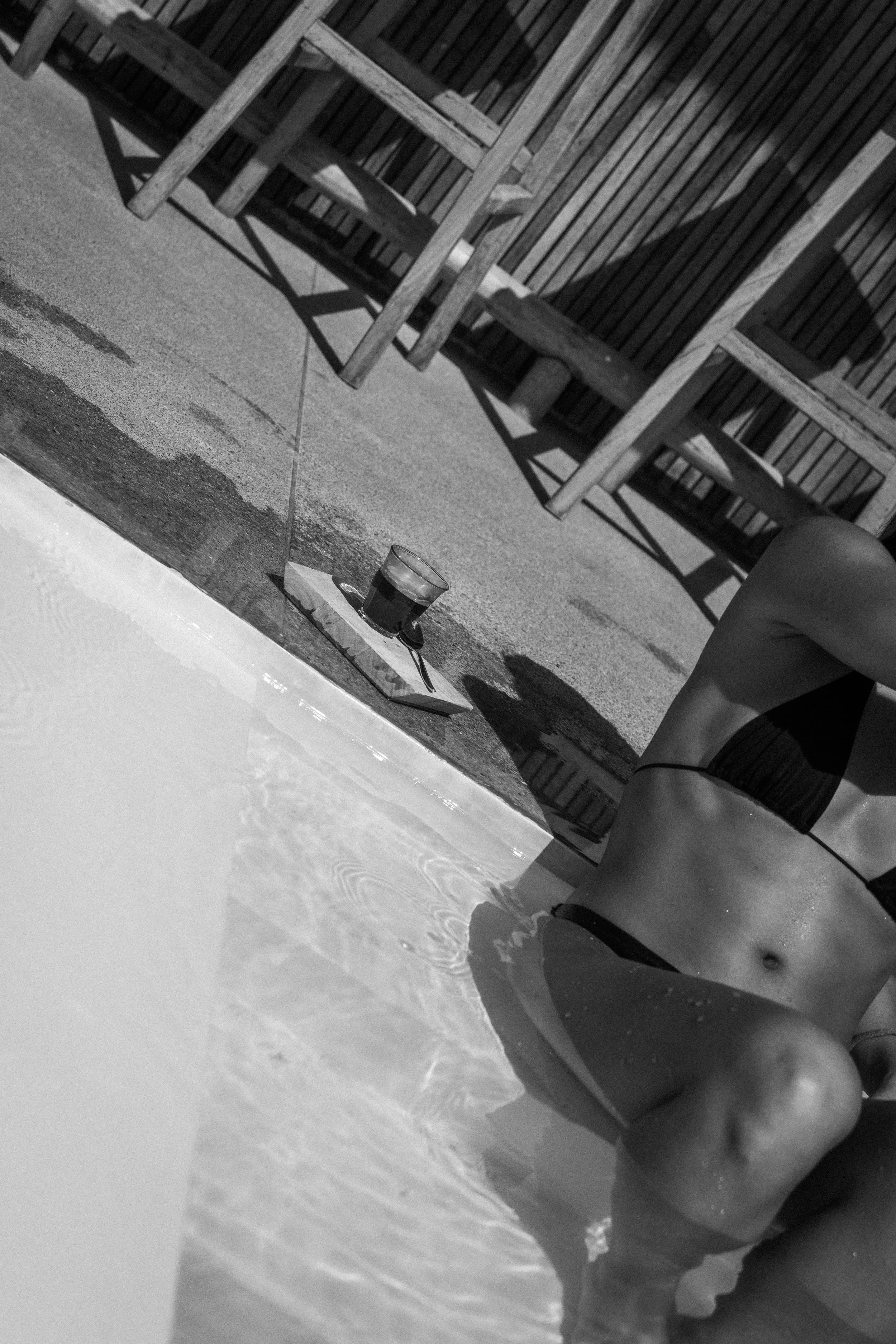 Schwarz-weiße Nahaufnahme eines nackten, jungen Mädchens im Pool, Blick auf ihren Bauch, mit einer Hand auf ihrem Bein. Im Hintergrund sind leere Holzstühle und ein Tisch mit einem Getränk. Der Himmel ist nicht sichtbar, und das Bild ist in Schwarz-W