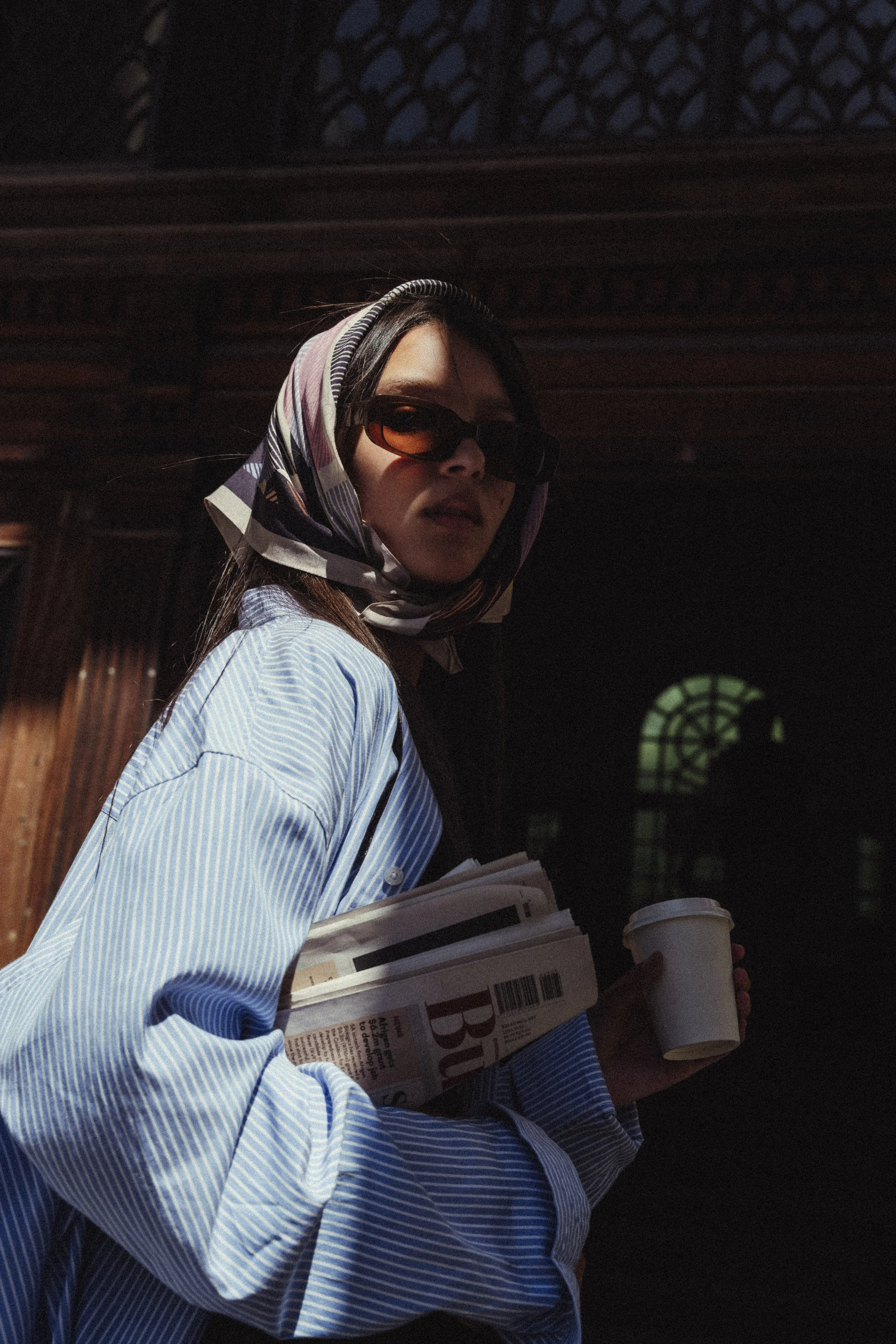 Junge Frau mit Sonnenbrille, Schal im Haar, blauer gestreifter Hemd, hält eine Tasse Kaffee und Zeitschriften. Im Hintergrund dunkles Holz mit Musterfenster. 
