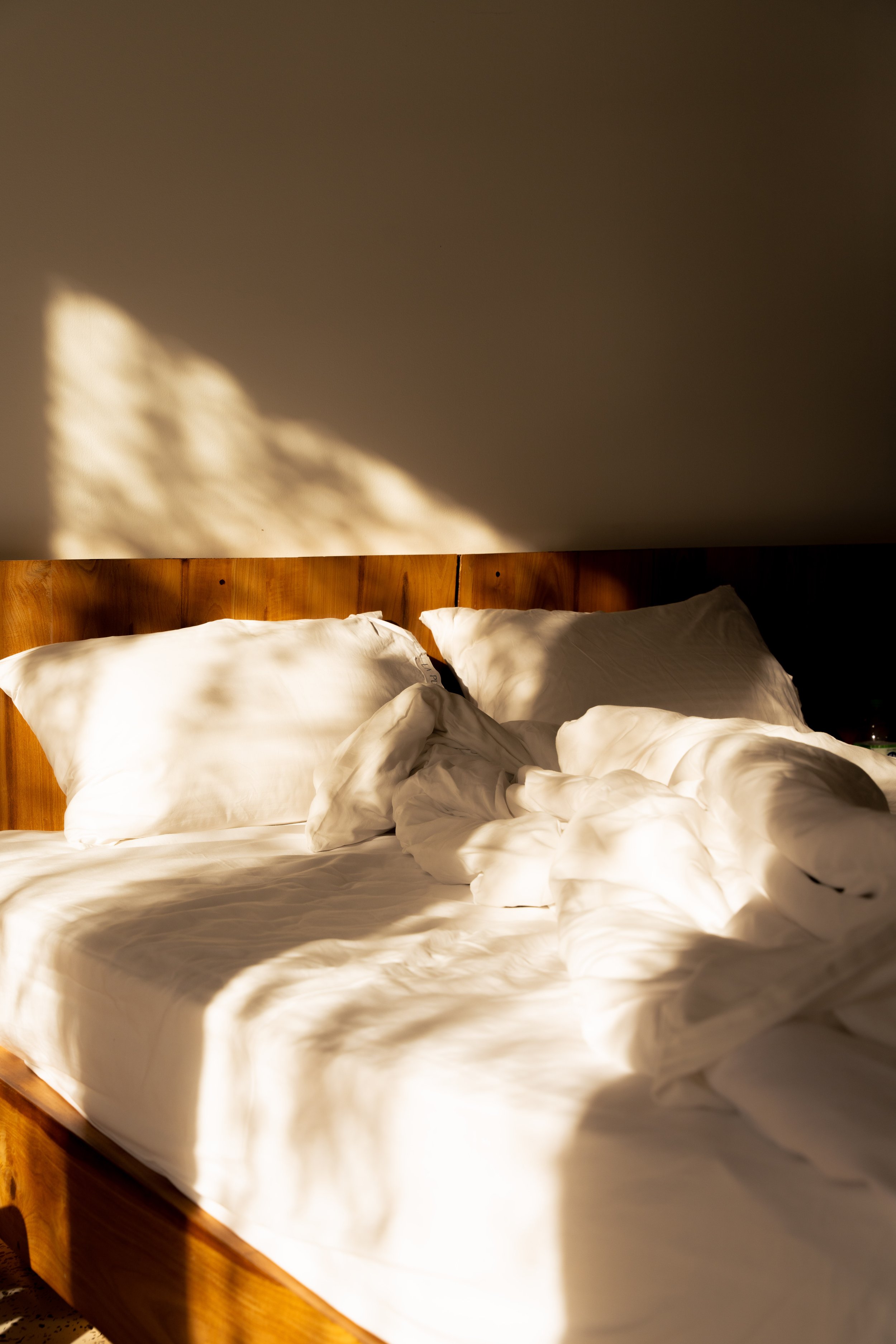 Verwehtes, unordentliches Bett mit weißen Laken und Kissen, beleuchtet durch Sonnenlicht, mit Schatten an der Wand im Hintergrund.
