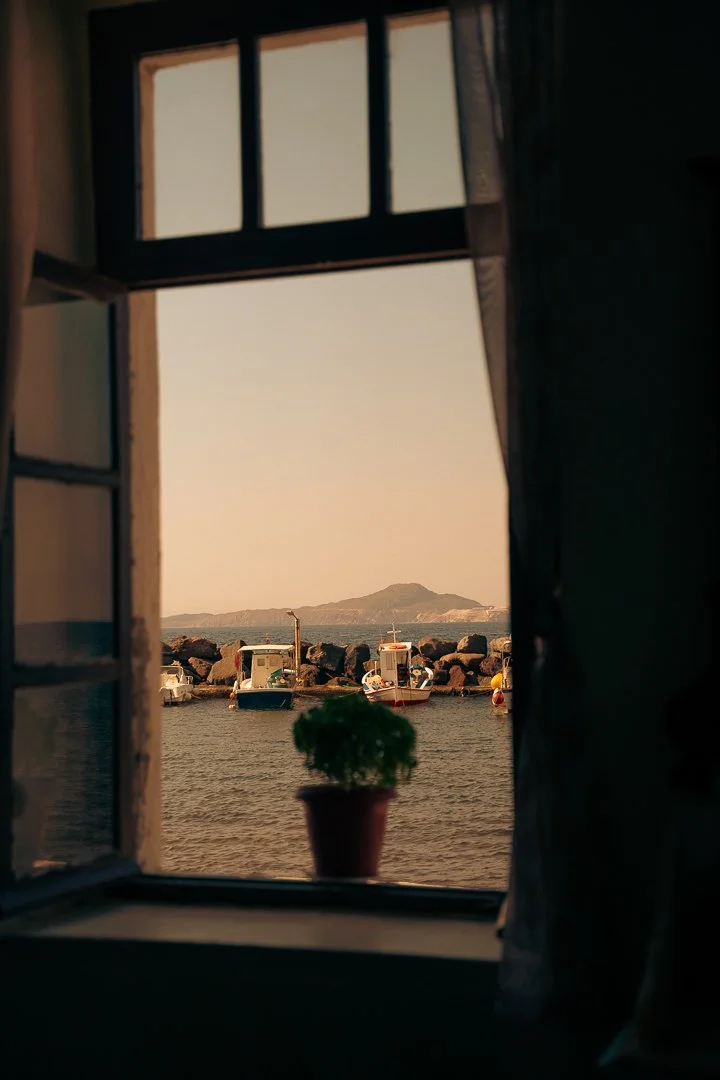 Ausblick durch ein Fenster auf Boote im Wasser mit einer kleinen Insel im Hintergrund, ein Blumentopf steht auf dem Fenstersims.