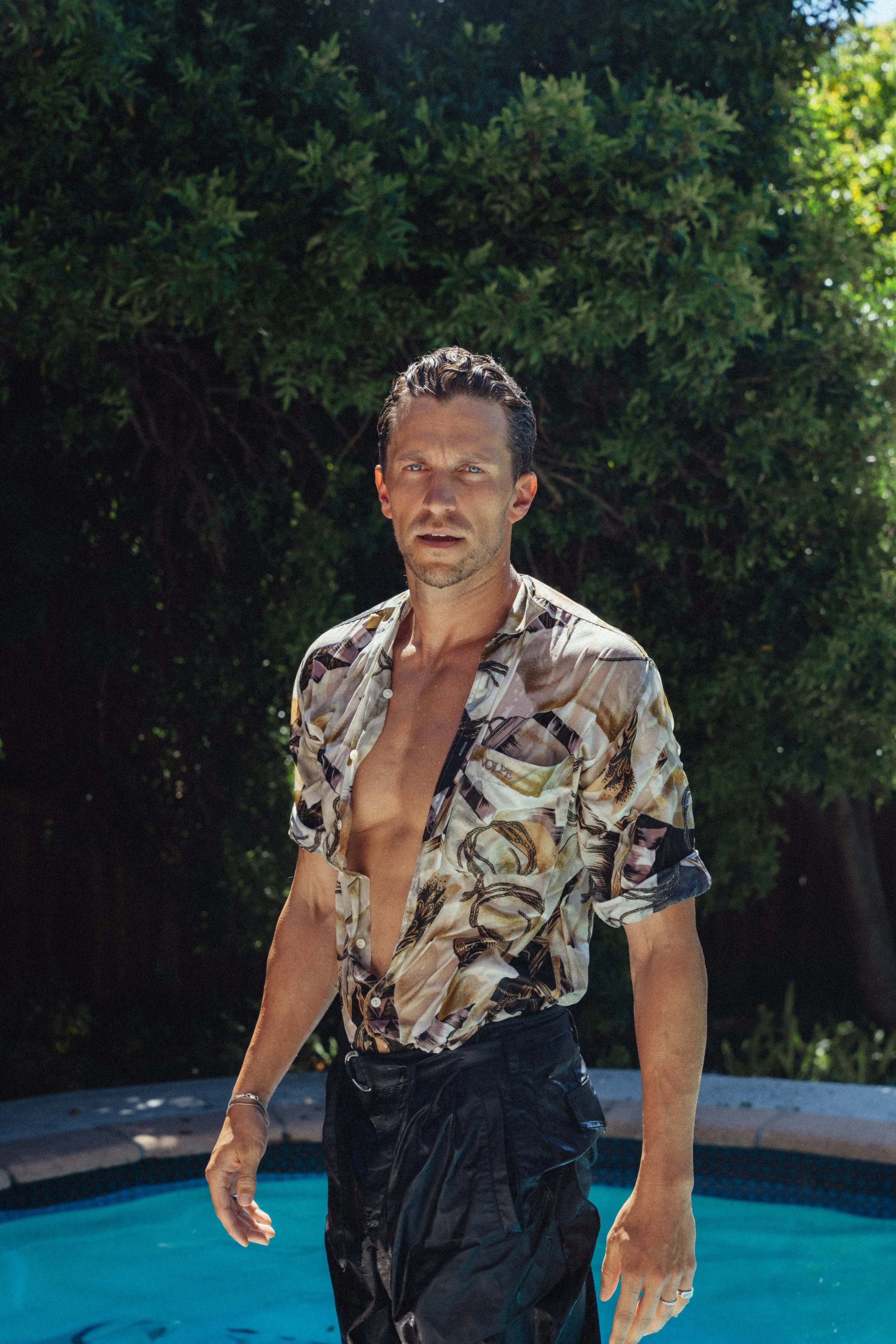 Ein Mann steht im Wasser vor einer grünen Baumkulisse, trägt ein offenes, gestreiftes Hemd und schwarze Schwimmhosen, blickt überrascht oder erstaunt.