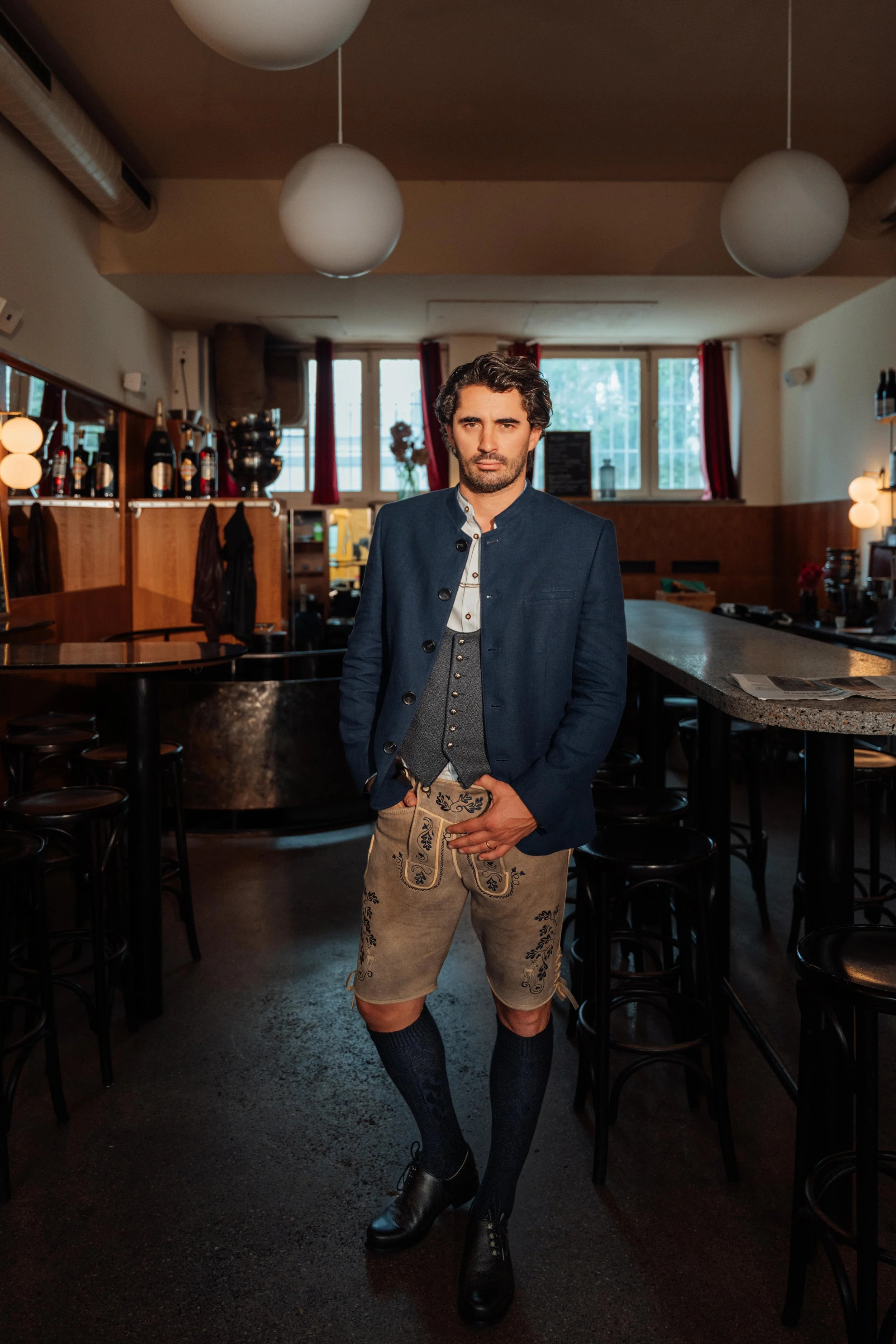 Mann in traditioneller bayerischer Kleidung steht in einer Bar, trägt eine blaue Jacke, eine Weste, Lederhosen, dunkle Socken und schwarze Schuhe.