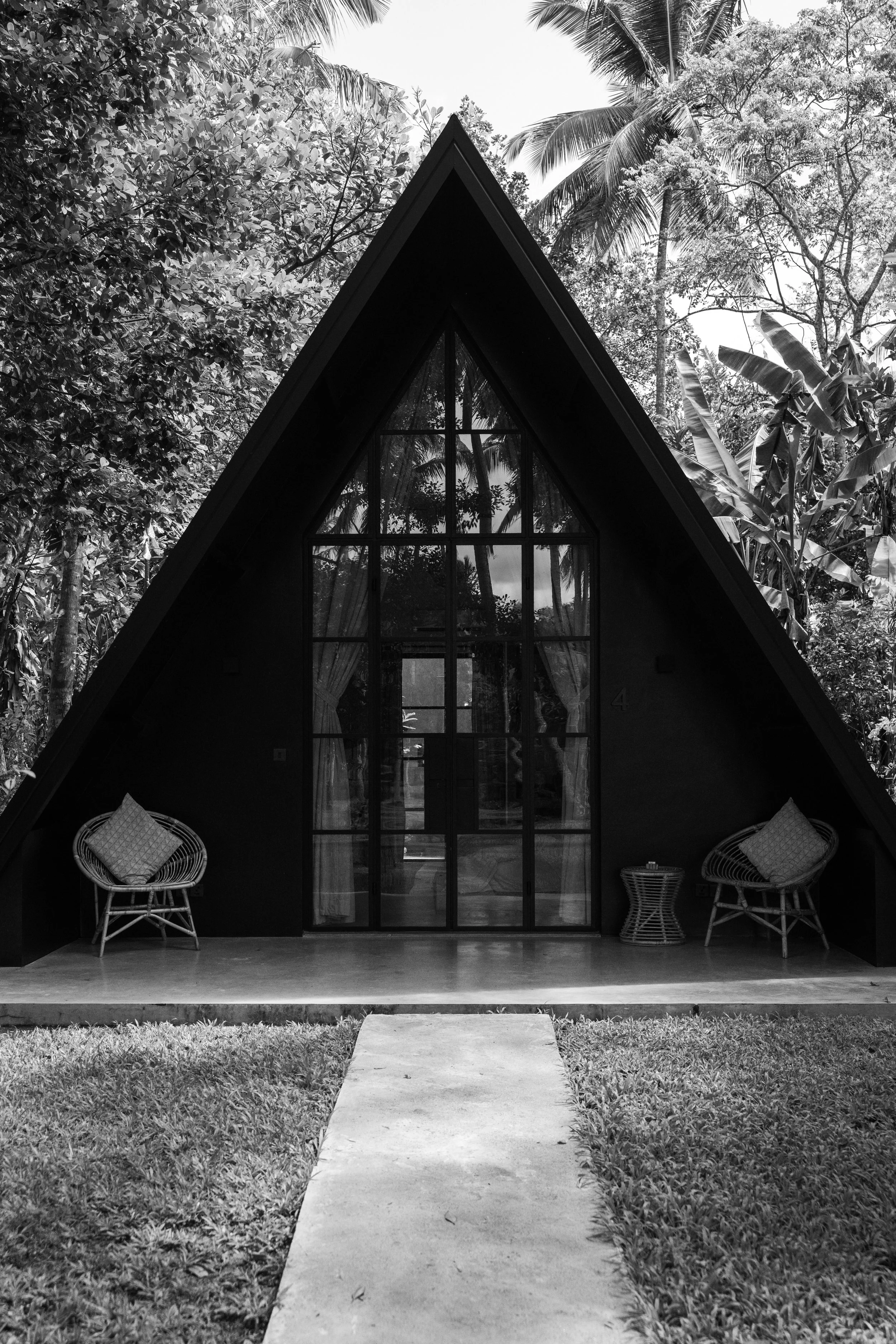 Ein modernes A-Frame-Haus mit großen Fensterfronten, umgeben von Bäumen, auf einer Wiese mit einem Gehweg davor, in Schwarz-Weiß.