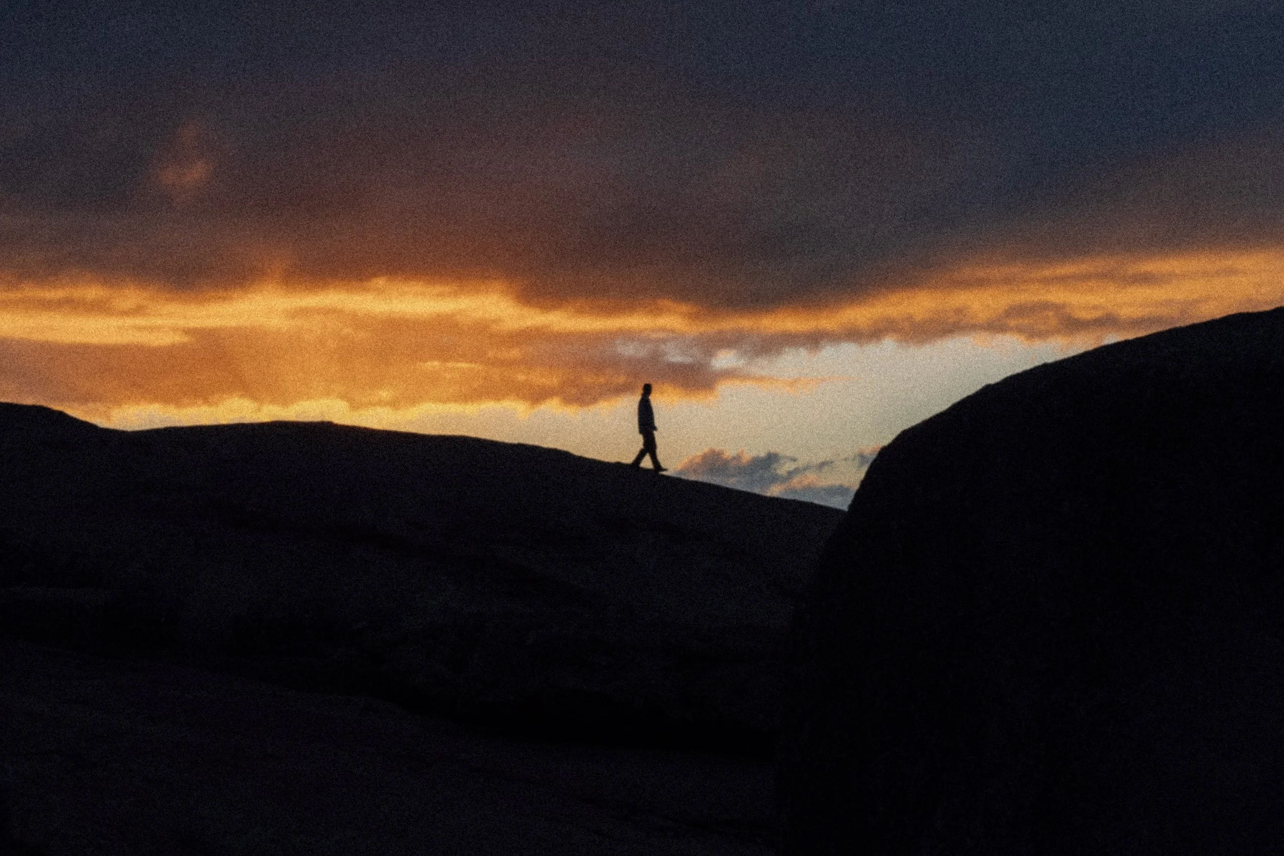 Schatten einer Person beim Spaziergang bei Sonnenuntergang auf einer hügeligen Landschaft mit dunkelblauem Himmel und orangefarbenen Wolken im Hintergrund.