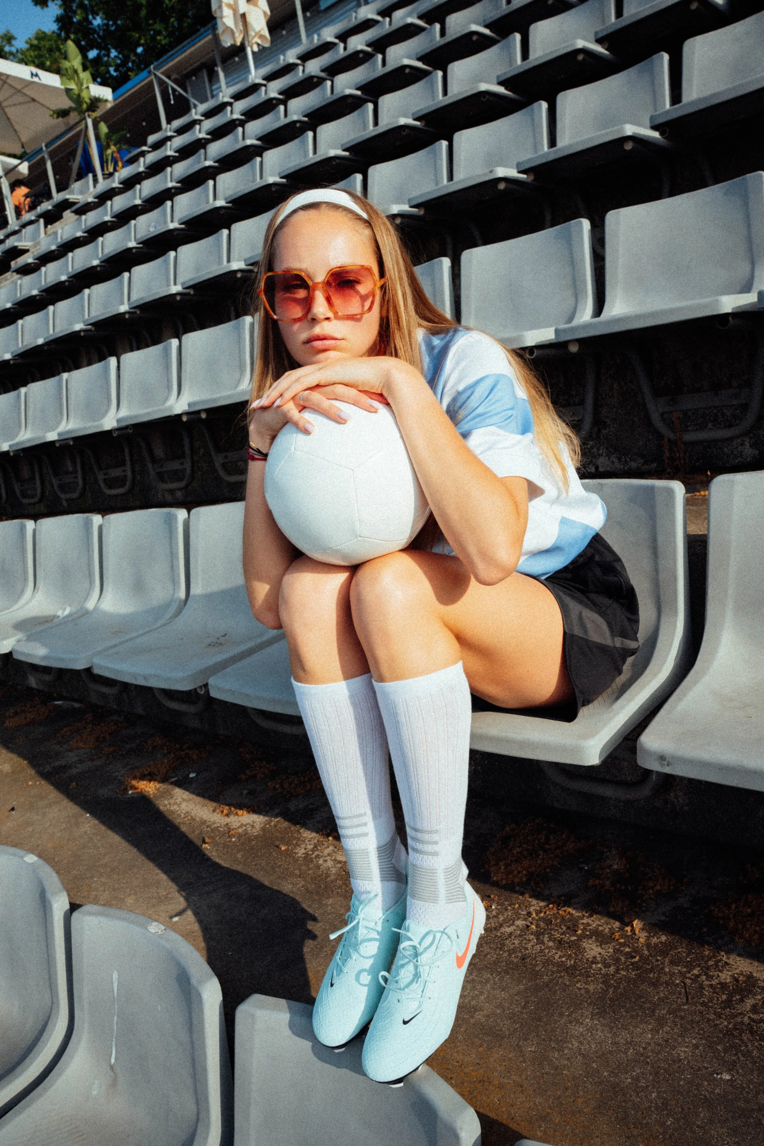 Junge Frau mit Sonnenbrille sitzt auf Sitzbank im Fußballstadion, hält einen Fußball, trägt Sportschuhe, hohe Socken, kurze Hosen und ein sportliches T-Shirt.