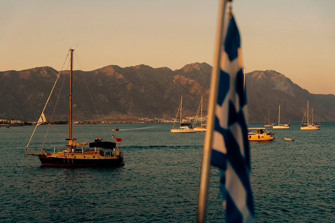 Segelboote auf dem Wasser mit Bergkulisse im Hintergrund, eine Griechenland-Flagge im Vordergrund